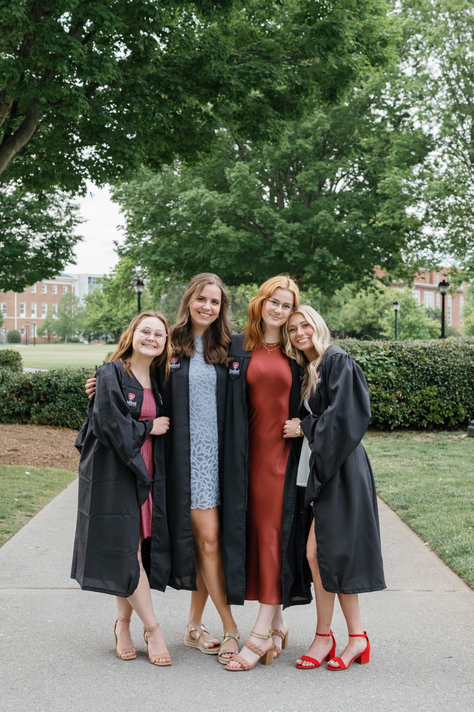 A group of four senior students standing together, smiling, and celebrating their graduation. Each wears a black graduation gown, with varying dress styles in different colors. They stand in front of lush green trees on Radford University's campus, exuding a joyful and proud moment.