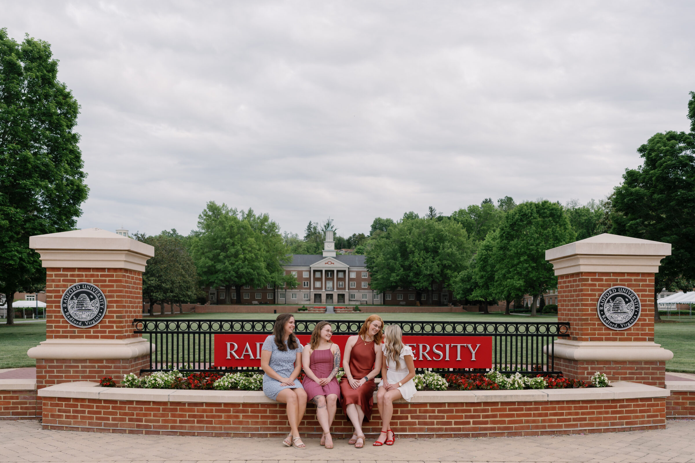 Four senior students sitting on a brick wall in front of the Radford University sign. They smile and share a joyful moment, dressed in colorful dresses and graduation gowns. The iconic university building and lush green trees are visible in the background, creating a beautiful and celebratory atmosphere.