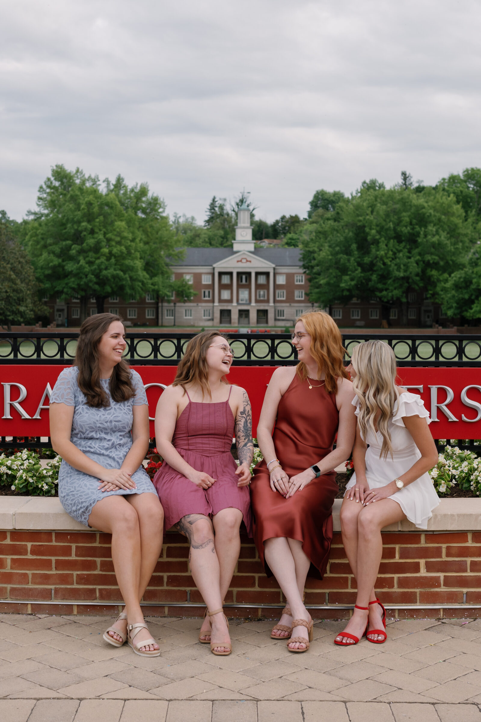 Four senior students sitting on a brick wall in front of the Radford University sign, laughing and enjoying the moment. The students, dressed in colorful dresses, smile and interact playfully with each other. The university’s iconic building and green landscape create a beautiful backdrop.