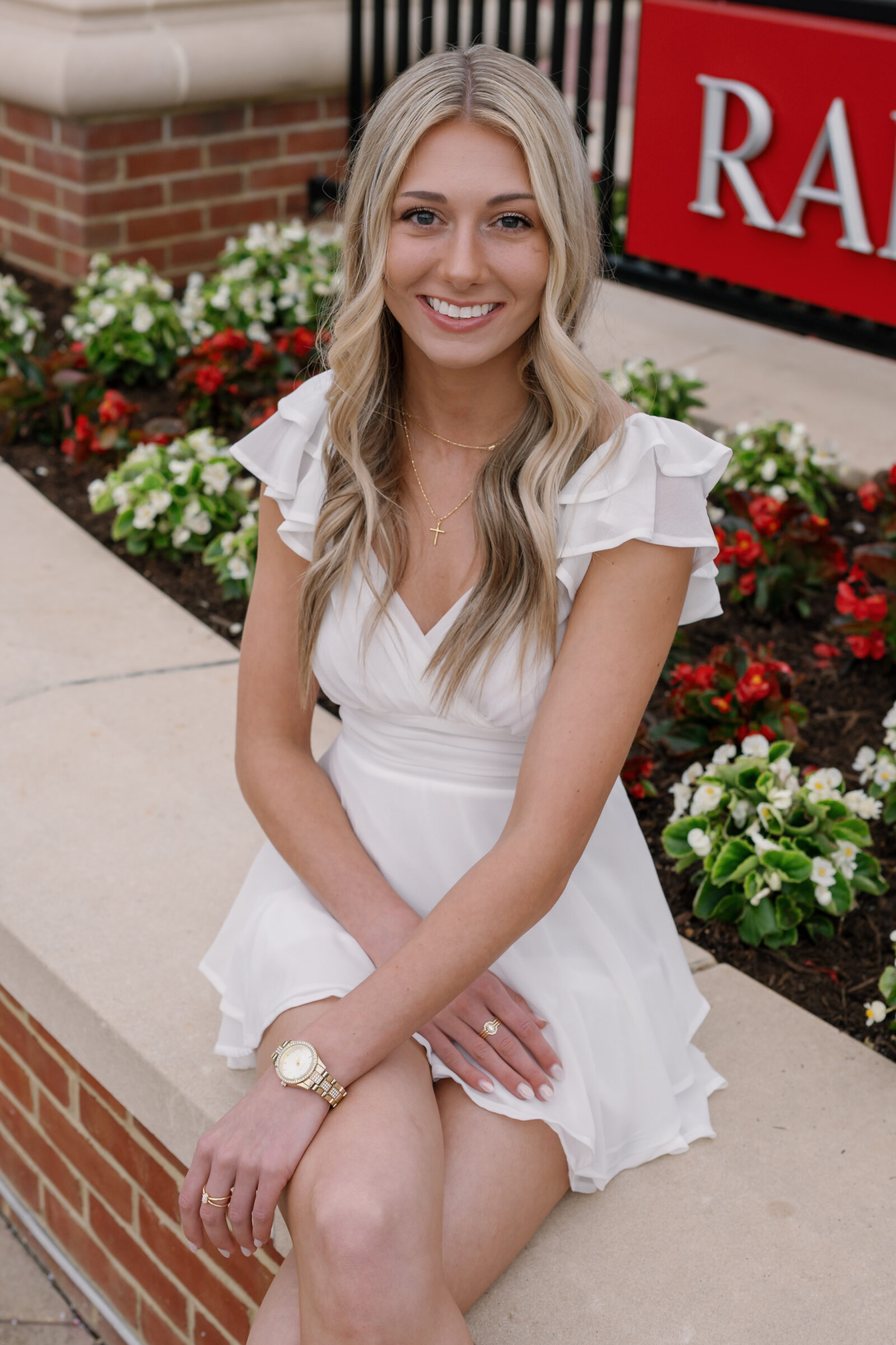 A close-up portrait of a senior student sitting on a stone bench in front of colorful flowers. She smiles warmly, wearing a white ruffled dress, gold jewelry, and a watch, with a Radford University sign visible in the background.