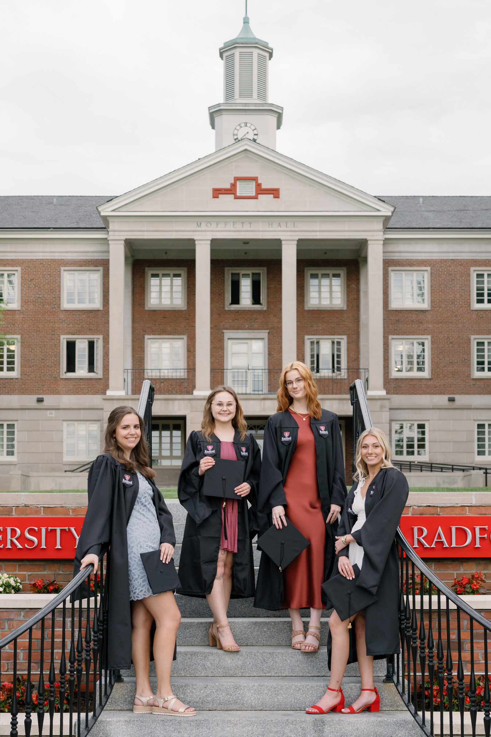 Four senior students in their graduation gowns posing together on the steps of Moffett Hall at Radford University. They each hold their graduation caps and smile proudly, with the university’s iconic brick building and flowers framing the scene.