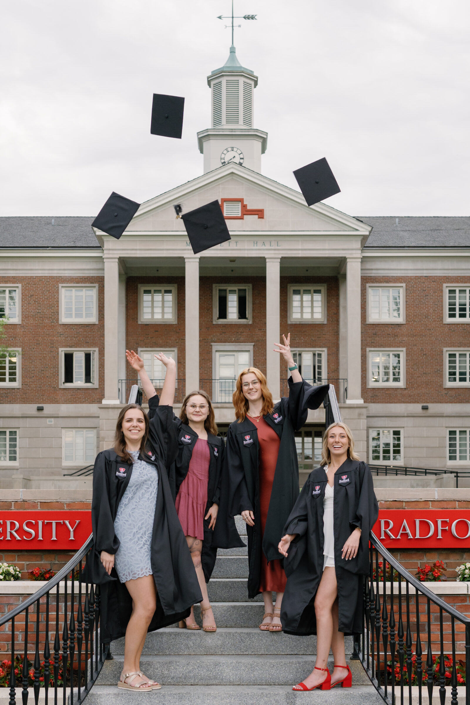 Four senior students celebrating their graduation by tossing their caps into the air in front of Moffett Hall at Radford University. They wear black graduation gowns and smile joyfully, with the university’s iconic building and the red Radford University sign in the background.