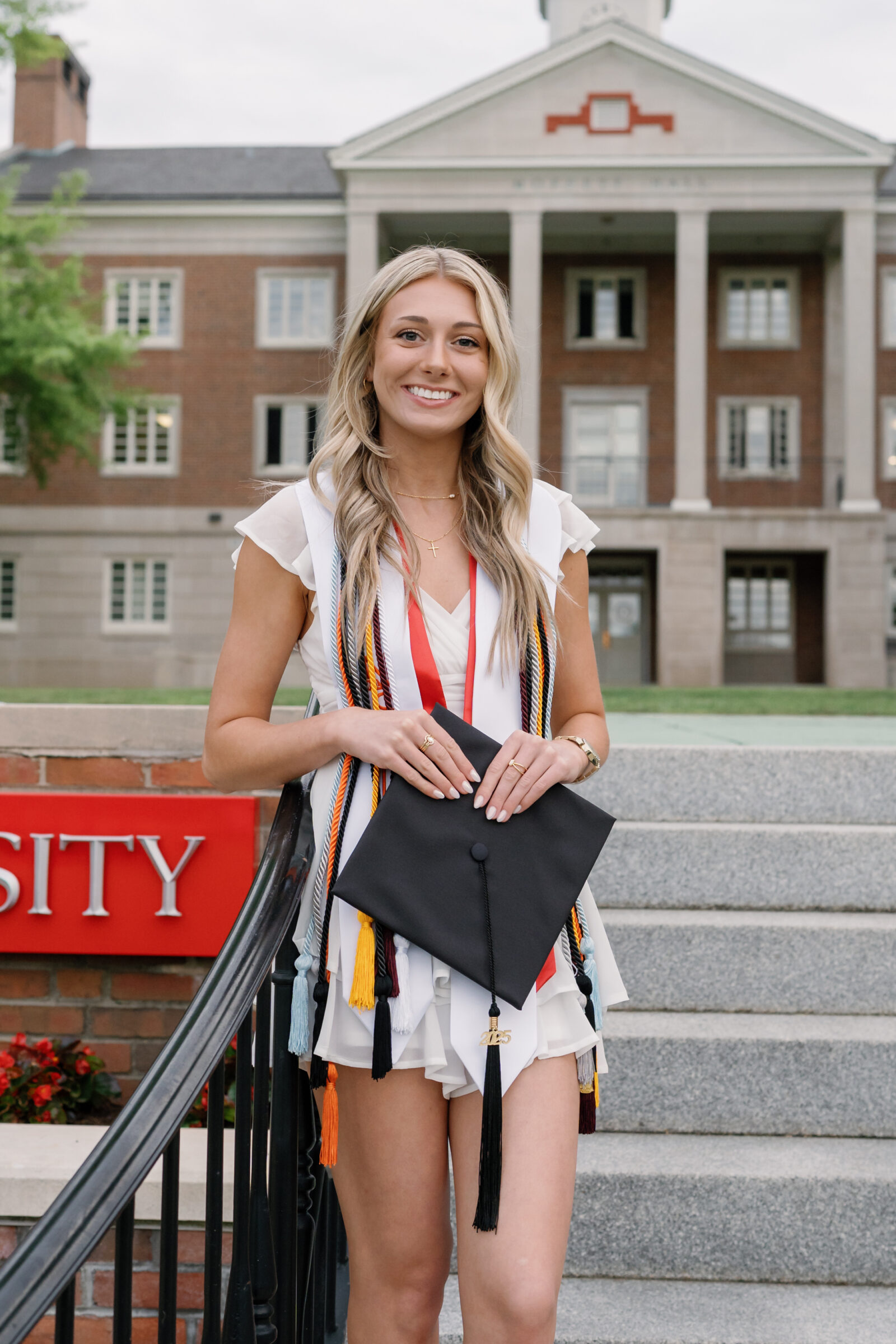 A senior student standing proudly in front of Moffett Hall at Radford University, wearing a white ruffled dress and holding her graduation cap adorned with colorful cords. She smiles at the camera, with the red Radford University sign and the iconic building in the background.