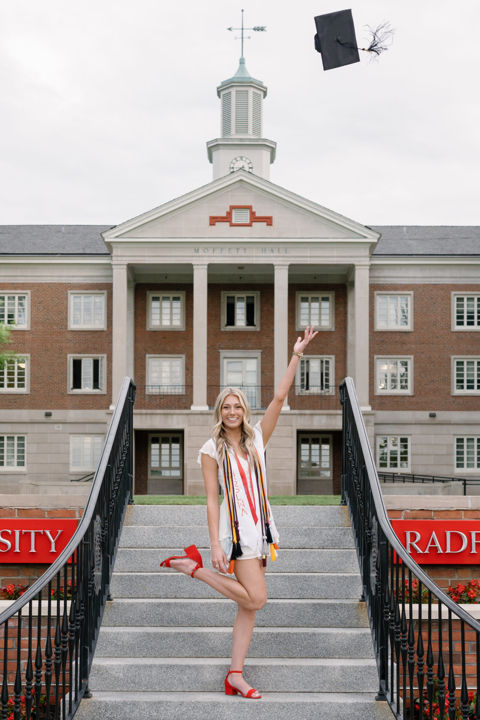 A senior student celebrating her graduation by tossing her cap into the air while standing on the steps of Radford University's Moffett Hall. She wears a white ruffled dress and colorful graduation cords, with red heels, smiling joyfully as her cap floats above her.