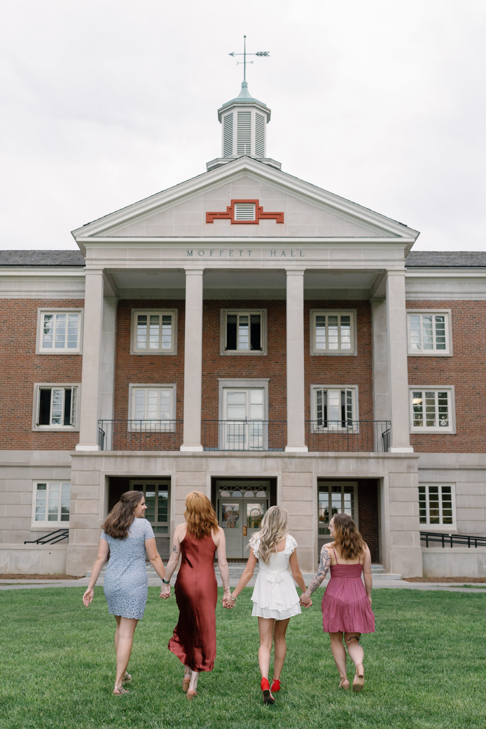Four senior students walking hand in hand across the grass in front of Moffett Hall at Radford University, dressed in colorful graduation dresses. The iconic brick building and columns provide a proud and celebratory backdrop.