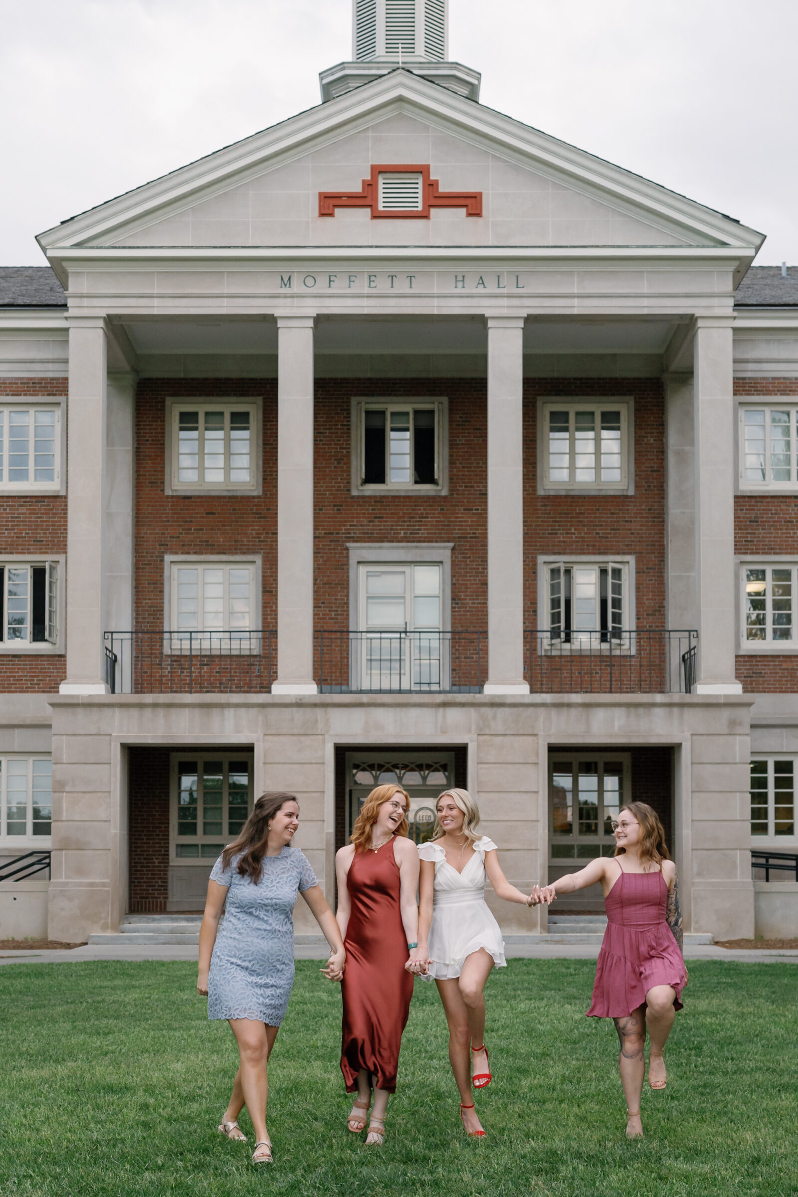 Four senior students holding hands and walking joyfully across the lawn in front of Moffett Hall at Radford University. They wear colorful dresses and graduation gowns, smiling and celebrating together, with the iconic building and green grass as the backdrop.
