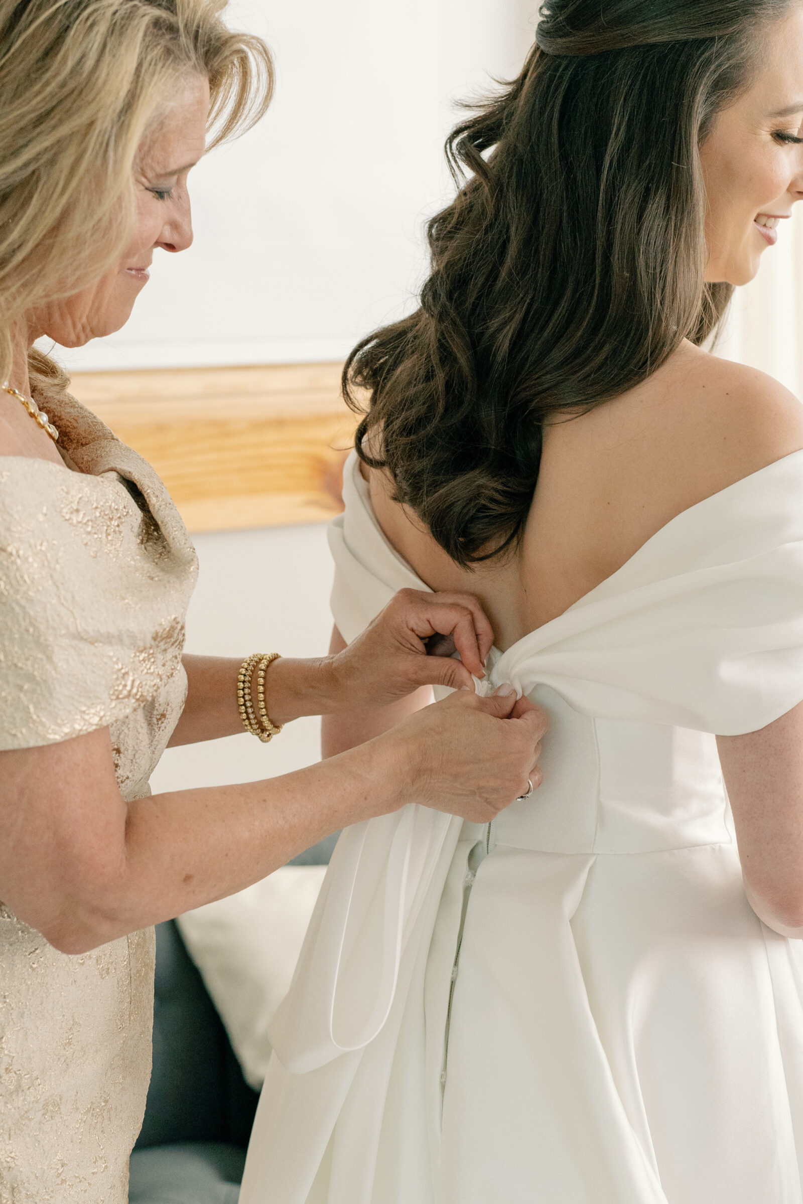 A heartfelt moment as the bride's mother helps her fasten the buttons of her wedding gown at Middleburg Barn. The soft, romantic light fills the room, highlighting the elegance of the off-shoulder gown and the bride's long, flowing hair. The intimate gesture captures a beautiful, timeless moment of support and love between mother and daughter, making this part of the wedding day truly special. The background is softly blurred, allowing the focus to remain on the bride and her mother.