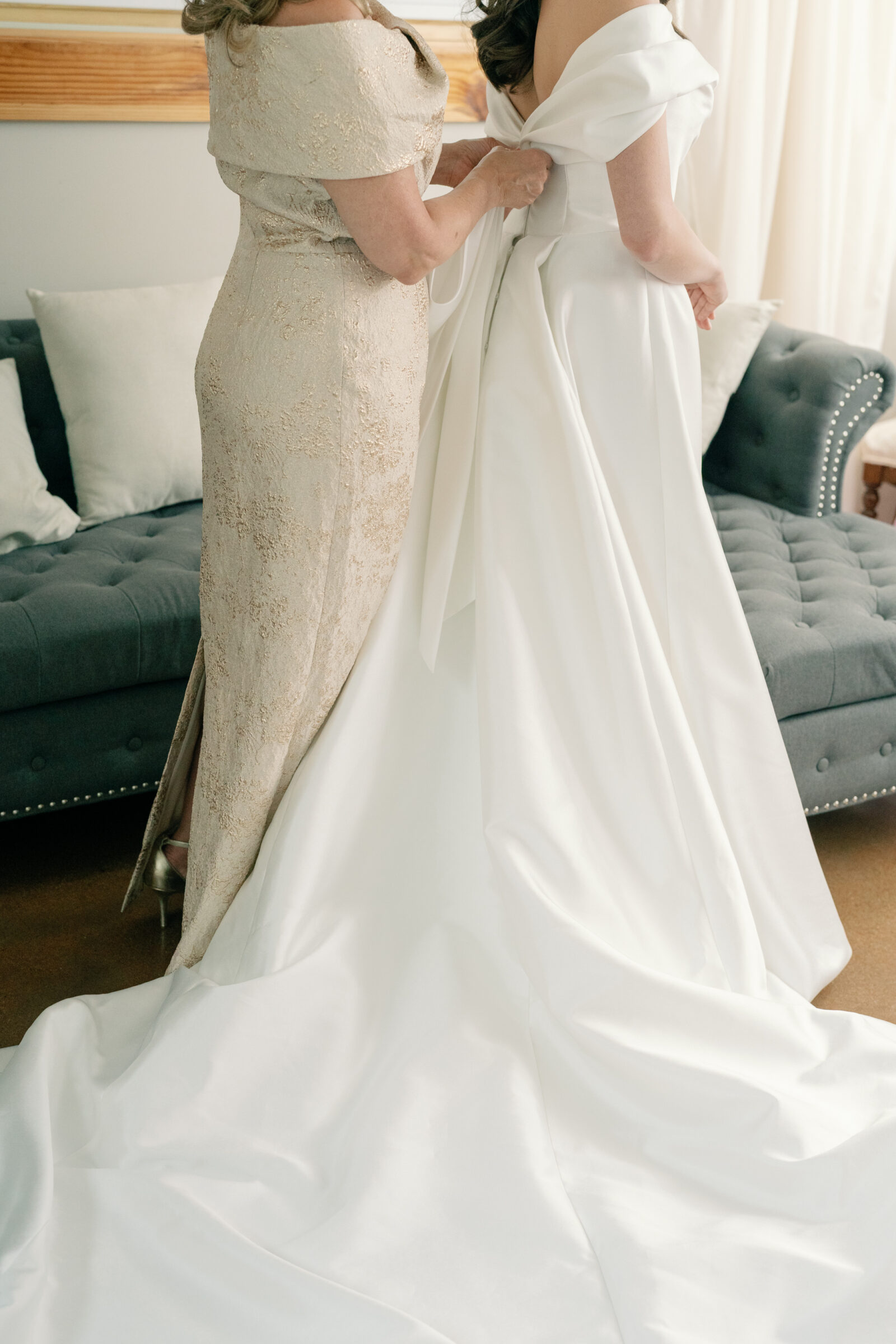 The bride’s mother lovingly adjusts the bride’s gown at Middleburg Barn, helping her prepare for the ceremony. The bride’s stunning off-shoulder dress, with a long, flowing train, is carefully smoothed out, showcasing the elegant satin fabric. The soft, golden tones of the mother’s dress and the cozy, well-lit room add to the intimate and emotional moment shared between the two before the big day.