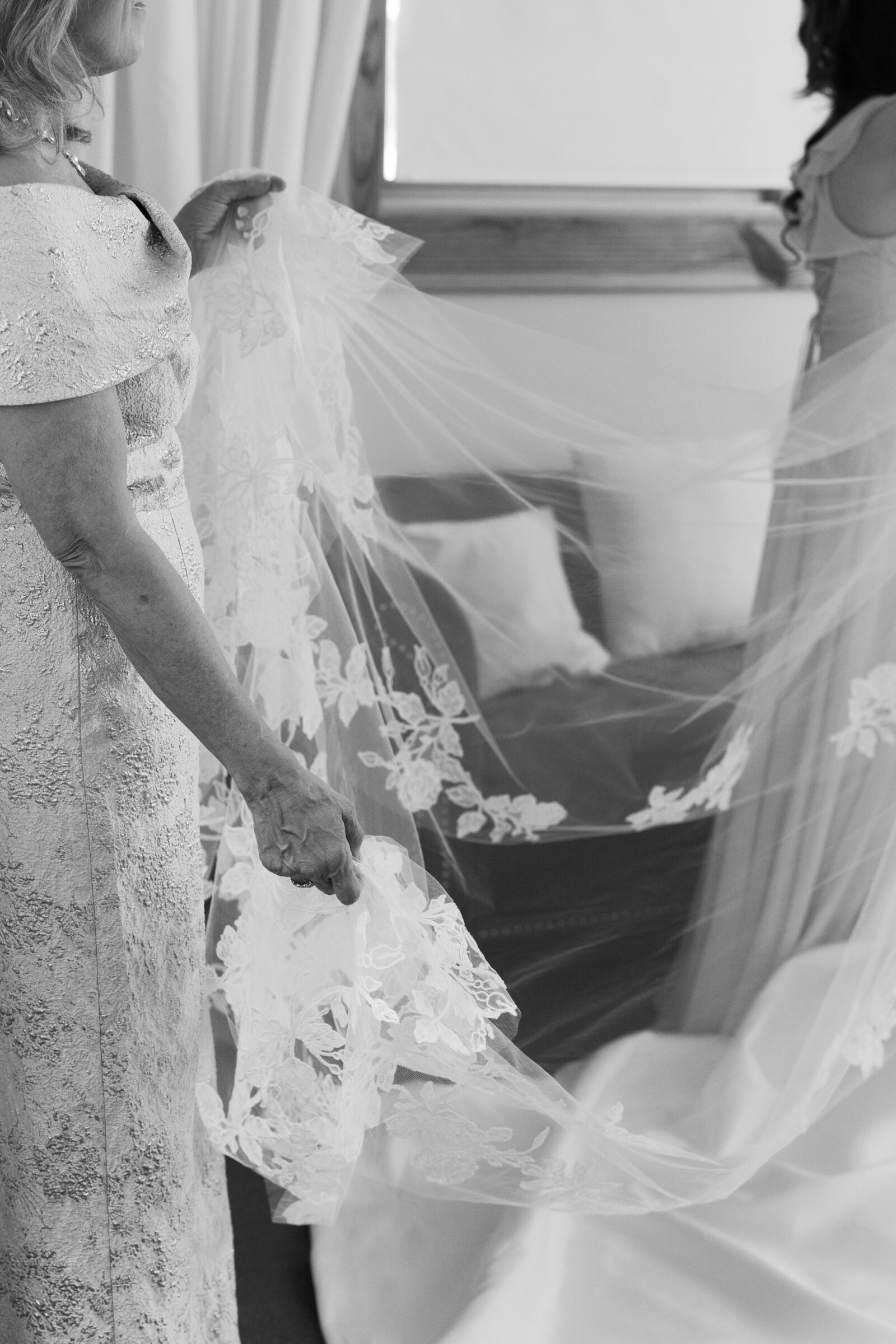 The bride’s mother tenderly holds the lace-edged veil as she prepares to place it on the bride, whose back is turned as a bridesmaid stands nearby. The veil, adorned with intricate floral embroidery, flows delicately through the air in a touching, pre-ceremony moment at Middleburg Barn. The soft, romantic atmosphere is captured as the bride’s mother lovingly assists with the final touches before the big moment.