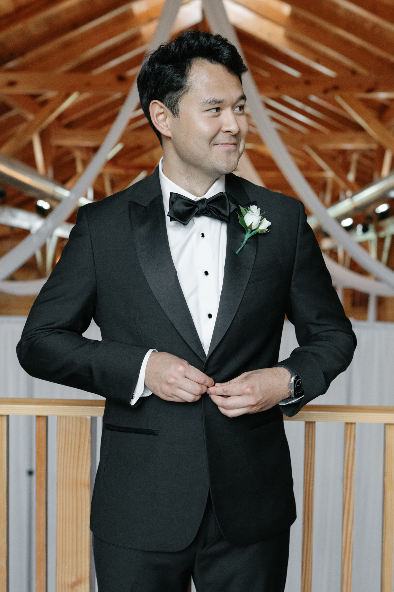 The groom stands in the barn at Middleburg Barn Wedding, adjusting his tuxedo jacket and smiling. He wears a classic black tuxedo with a black bow tie and a white rose boutonnière, ready for the ceremony.