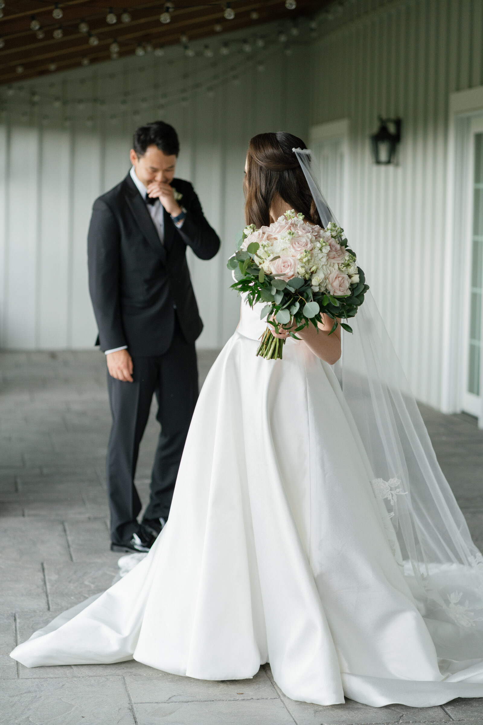 The bride, dressed in an elegant off-the-shoulder wedding gown, stands with her back to the groom in front of Middleburg Barn. She holds a lush bouquet of roses and greenery while the groom wipes a tear from his eye, moved by the emotional first look.