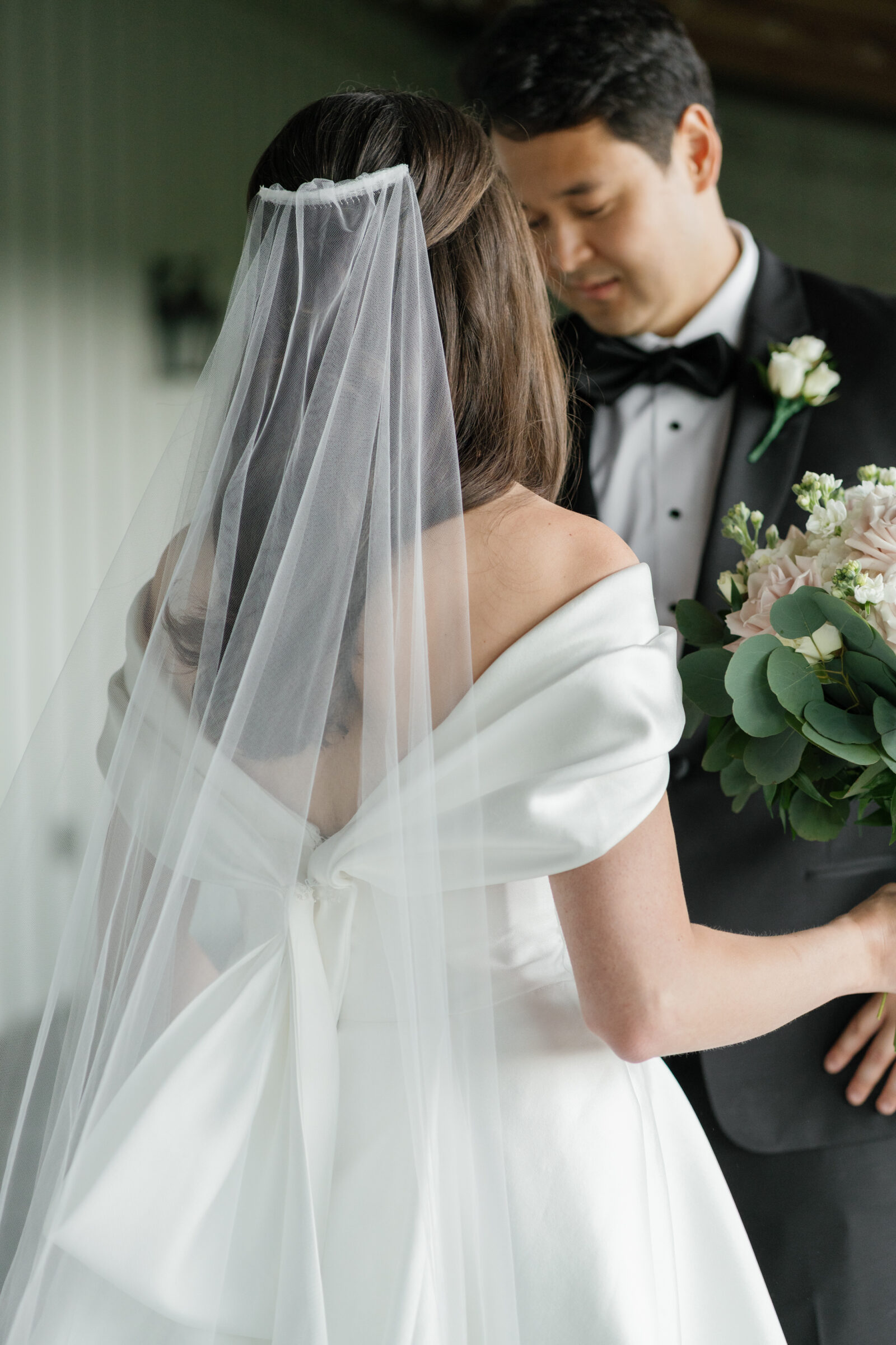 The bride in her off-the-shoulder wedding gown with a delicate veil stands close to her groom, who lovingly gazes at her while she holds her romantic bouquet. The soft fabric of the gown and veil frame the intimate moment at Middleburg Barn during their first look.