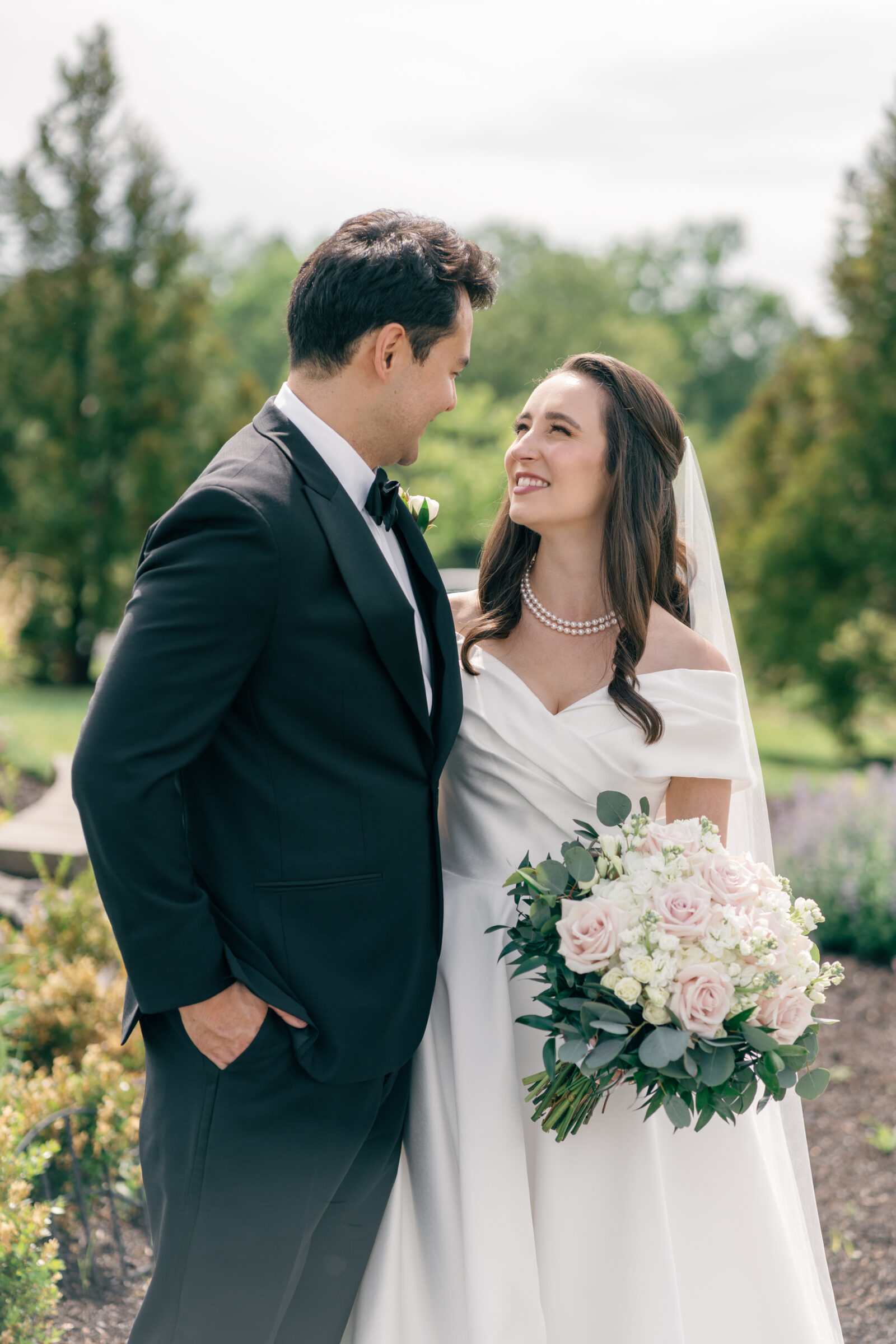 The bride and groom share a joyful moment as they smile at each other, with the bride holding her lush bouquet of pink roses and greenery. The couple is framed by the vibrant outdoor scenery of Middleburg Barn, capturing the essence of their love and happiness on their wedding day.