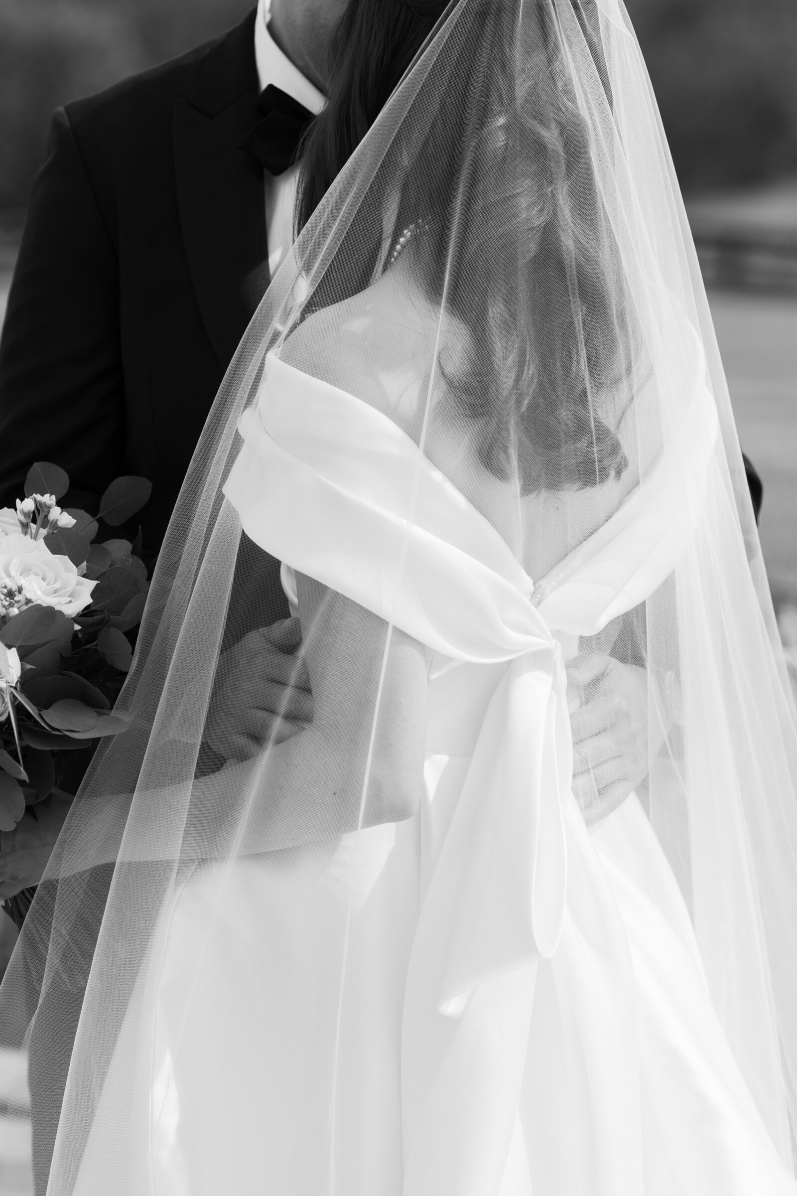 A romantic close-up of the couple embracing, with the bride's elegant gown and veil flowing delicately around her. Her bouquet of soft pink roses and greenery rests in the groom's hand as he gently holds her waist, creating a moment of intimate connection amidst the serene outdoor backdrop.