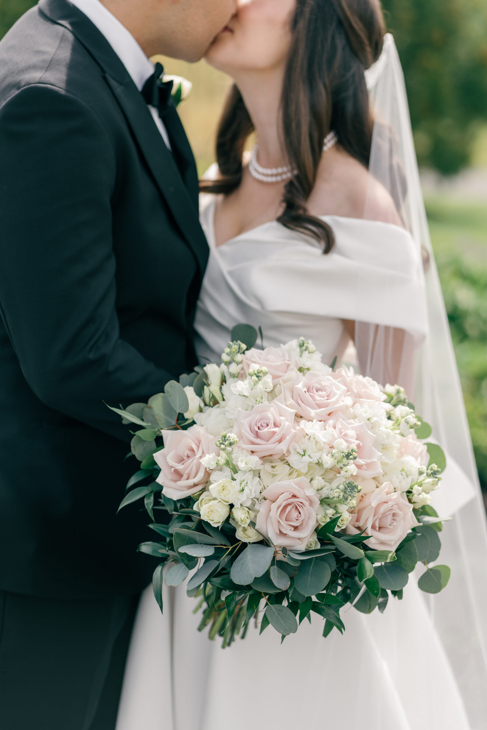 The bride and groom share a tender kiss as the bride holds her stunning bouquet of blush pink roses, white flowers, and lush greenery. The elegant gown and soft veil complement the romantic moment captured in the open air at Middleburg Barn.