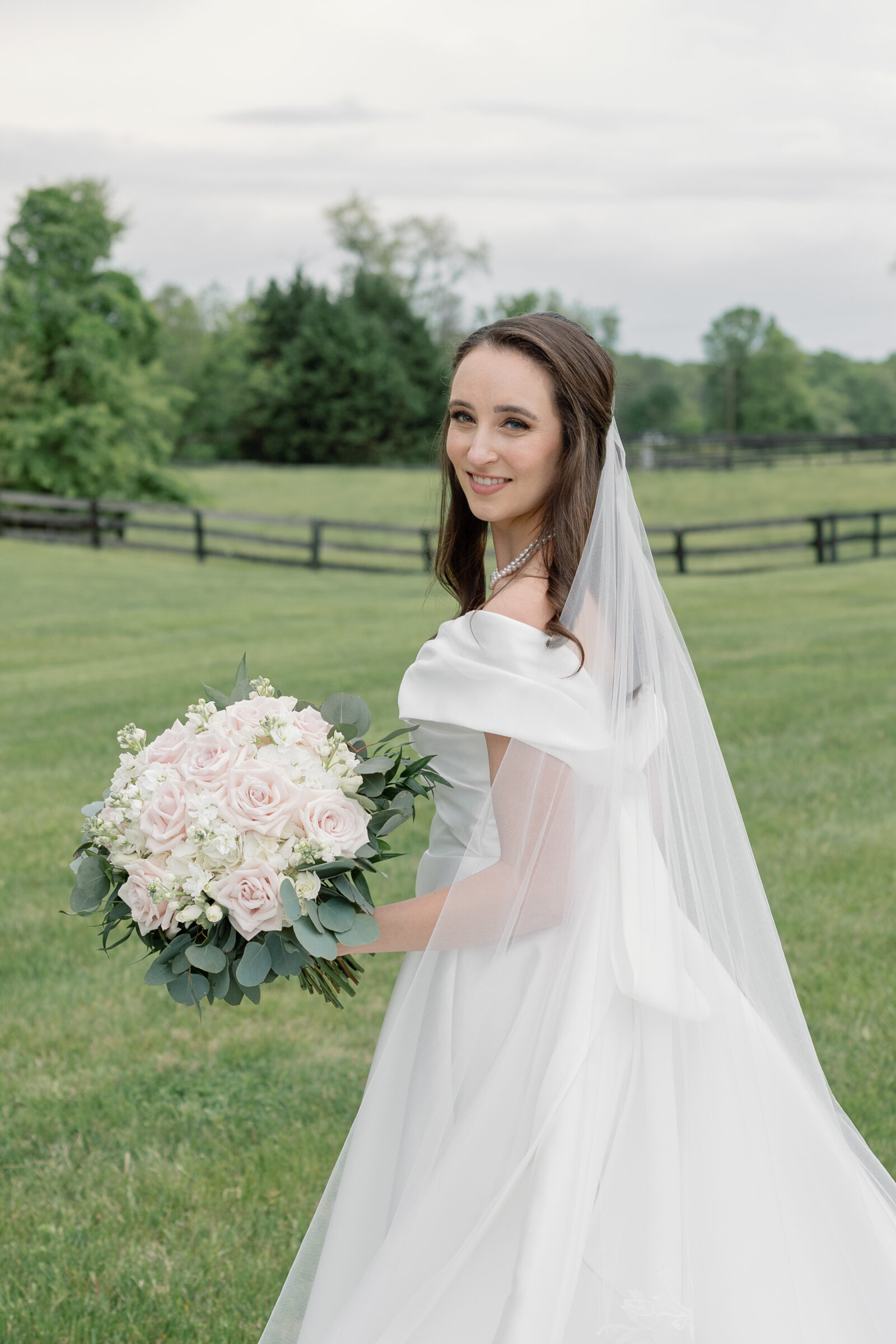 The bride beams with happiness as she holds her lush bouquet of pink roses and greenery, standing in the beautiful outdoor setting of Middleburg Barn. Her elegant white gown flows gracefully, and her veil catches the light as she smiles at the camera, showcasing the peaceful, picturesque backdrop of rolling green fields and fences.