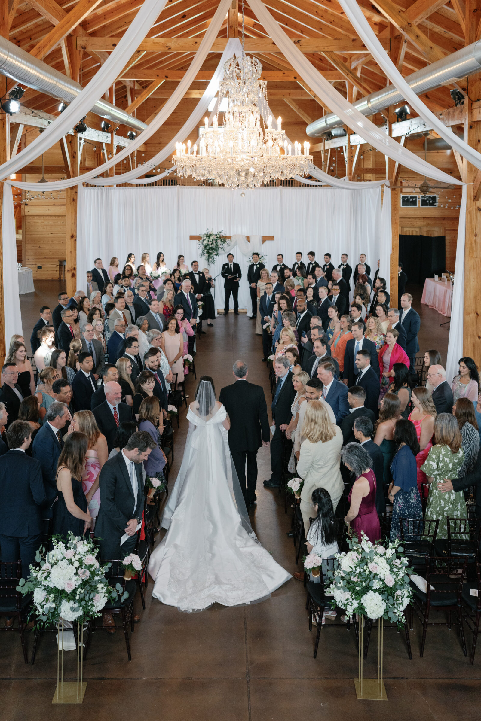 A wide shot of the wedding ceremony at Middleburg Barn, capturing the bride walking down the aisle with her father. The guests are seated on either side, surrounded by elegant floral arrangements and drapery hanging from the barn's wooden beams. The chandelier above adds to the grandeur of the setting, creating a beautiful, timeless moment.