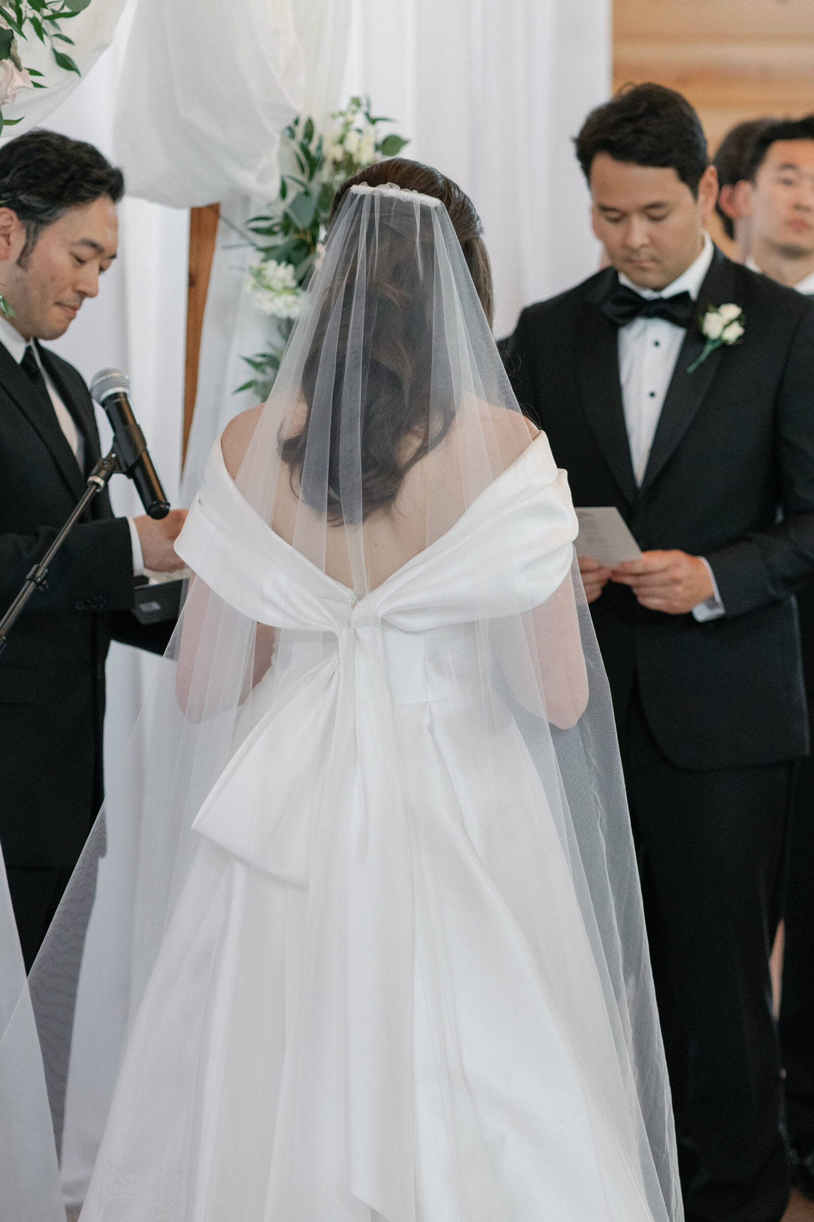 A close-up shot of the bride and groom during their ceremony at Middleburg Barn. The bride is standing with her back to the camera, her white veil flowing down her back, and her dress featuring an elegant bow detail at the waist. The groom, in a black tuxedo, is standing beside her, holding a card as they exchange vows, while a officiant stands in the background with a microphone.