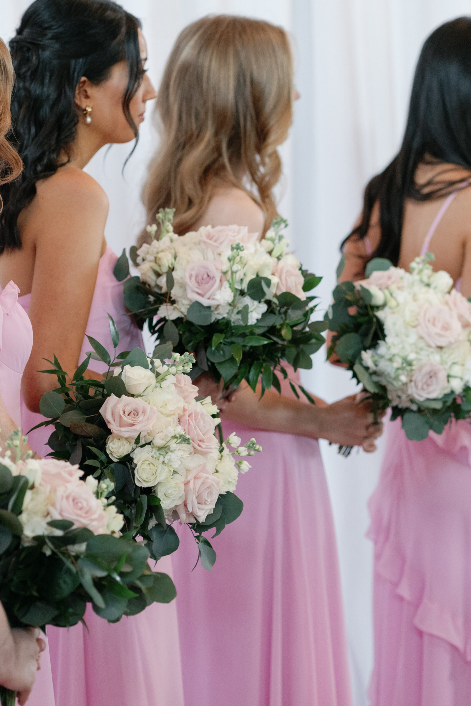 A close-up shot of the bridesmaids during the ceremony at Middleburg Barn. The focus is on the bridesmaids' bouquets, which are composed of blush pink roses, white flowers, and lush greenery. They are dressed in soft pink gowns, with the camera capturing them from behind as they stand in a row, watching the couple exchange vows.