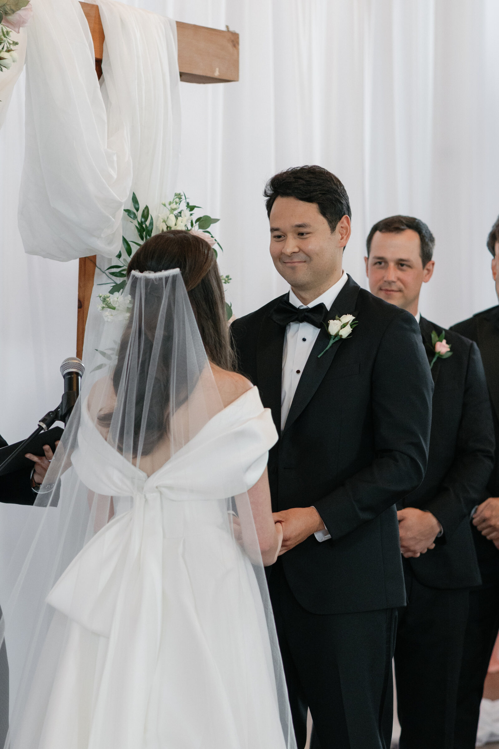 The bride and groom are holding hands at the altar during their wedding ceremony at Middleburg Barn. The bride, seen from behind, is wearing a stunning off-the-shoulder gown with a large bow at the back, and her veil gently flows as they exchange vows. The groom is smiling at the bride as he stands in a tuxedo, with the bridal party visible in the background.