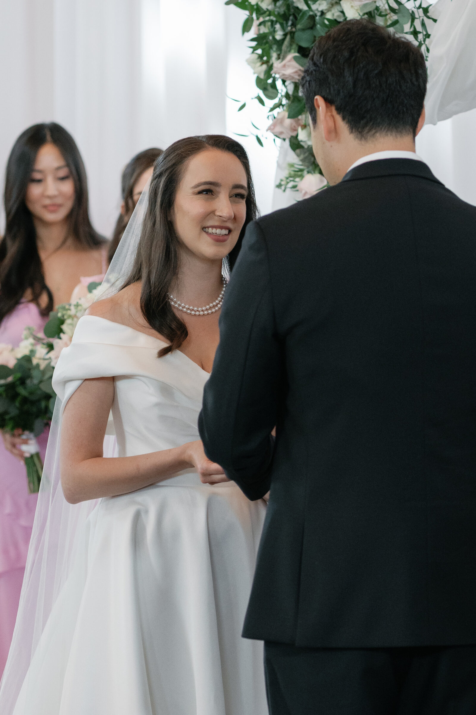 The bride and groom exchange smiles during their wedding ceremony at Middleburg Barn. The bride, wearing a classic off-the-shoulder gown with a veil, beams with joy as she holds the groom’s hands. The bridal party, dressed in soft pink dresses, stands in the background, holding bouquets.