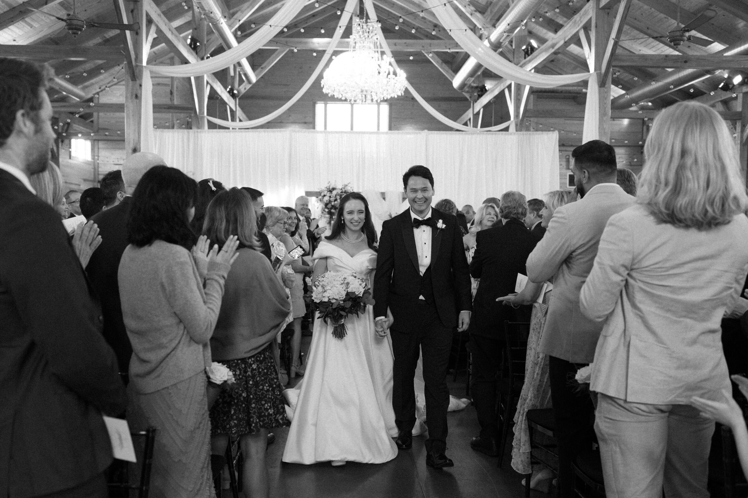 The newlyweds smile as they walk down the aisle, hand in hand, after their wedding ceremony at Middleburg Barn. Surrounded by guests clapping and cheering, the bride holds her bouquet, and the groom walks beside her, beaming with joy. The elegant venue features a grand chandelier and soft draping in the background.