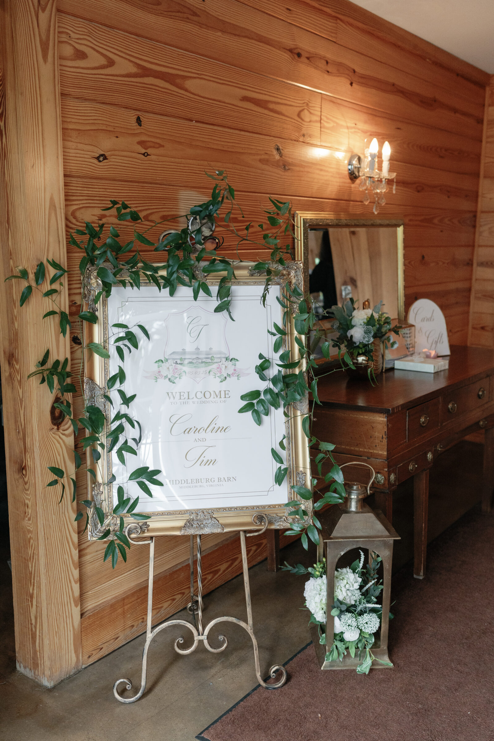 A vintage-inspired welcome sign adorned with lush greenery sits elegantly at the entrance of Middleburg Barn. The framed sign welcomes guests to the wedding of Caroline and Tim, with soft floral details and surrounding decor including a lantern and a small floral arrangement on a nearby wooden table.