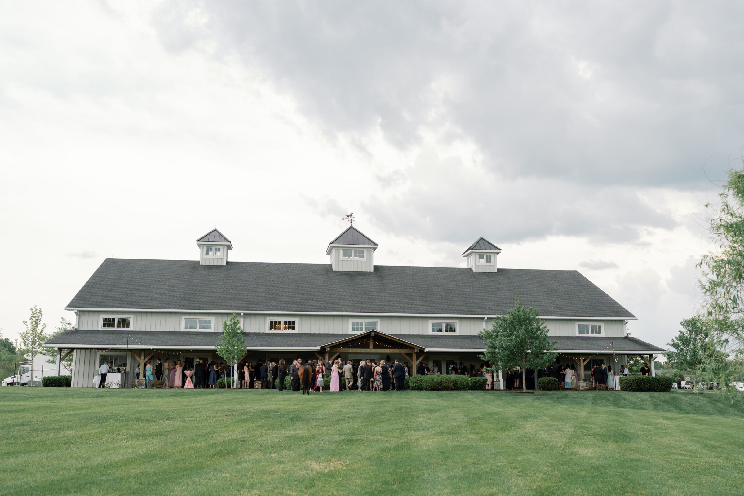 A wide shot of the Middleburg Barn venue with guests gathered outside. The barn’s large structure, with its white siding and charming cupolas, serves as the backdrop. Guests in formal attire mingle under a covered porch area, and the green grass and trees add to the picturesque rural setting.
