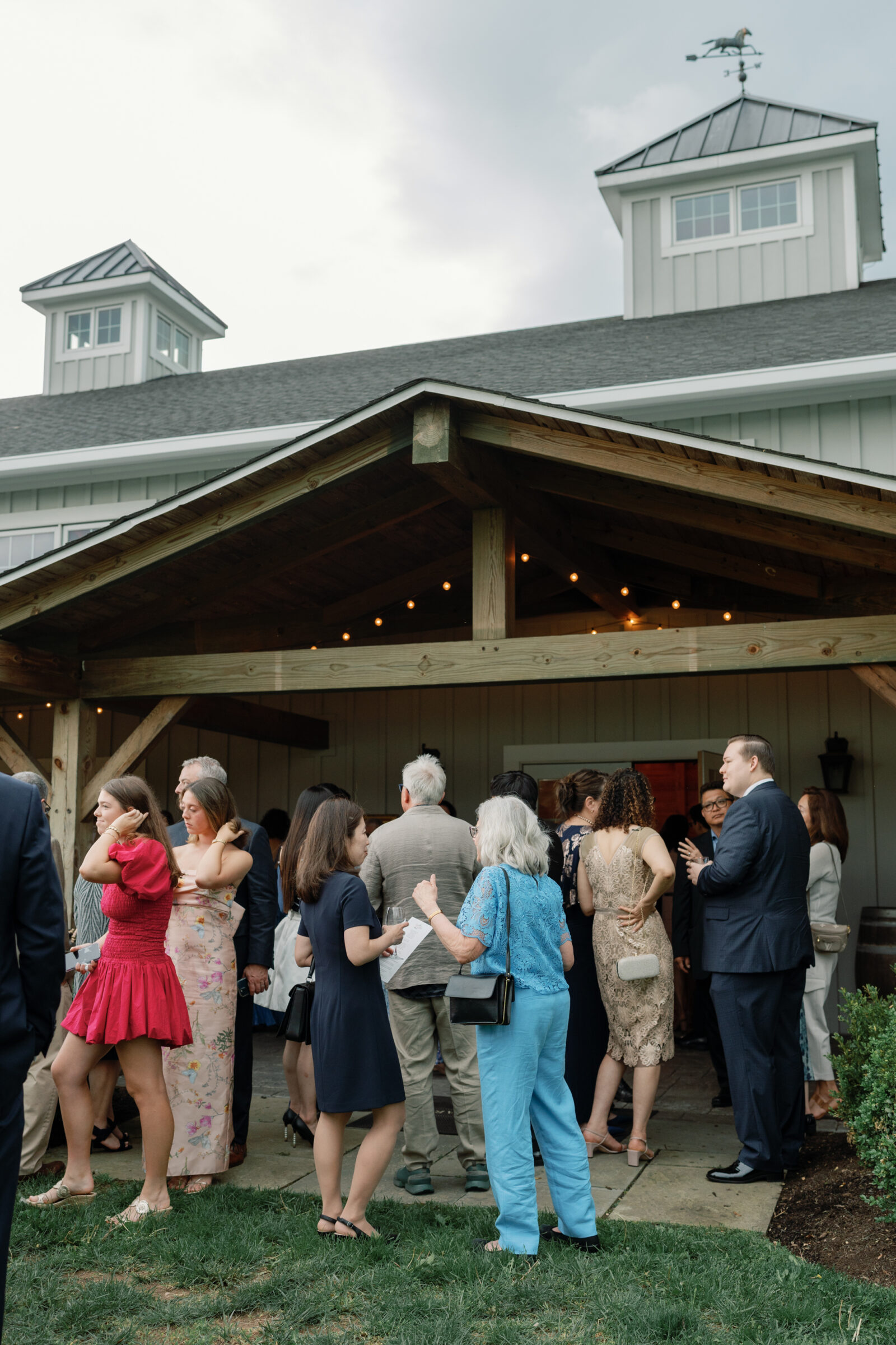A group of wedding guests gathered outside under a canopy at Middleburg Barn. The scene shows a variety of guests interacting and enjoying the reception. Some are wearing formal wedding attire, including dresses in shades of red, floral patterns, and suits. The barn is visible in the background with its wooden beams and string lights.
