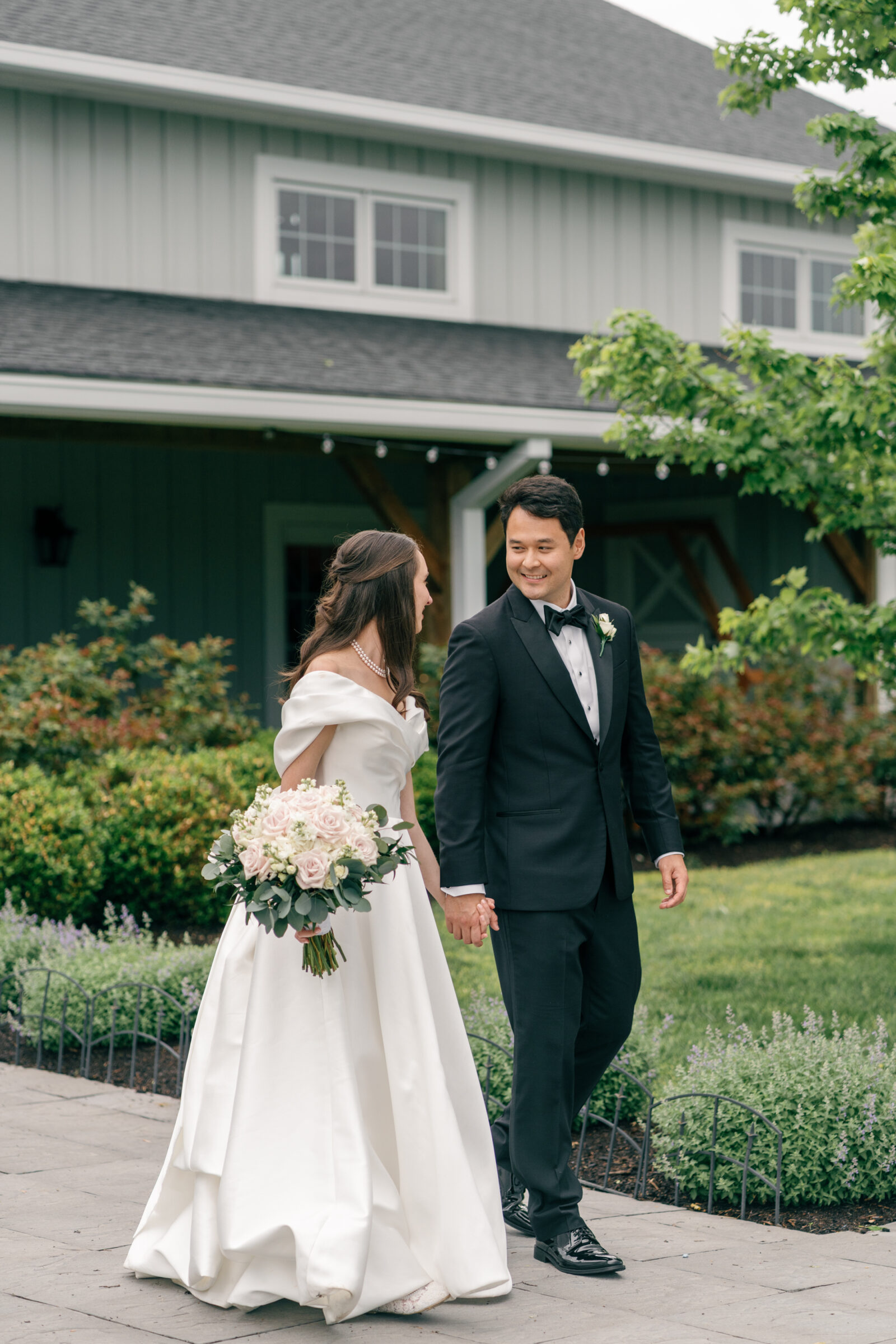 A bride and groom walk hand-in-hand through the grounds of Middleburg Barn, smiling and looking at each other. The bride wears a satin off-the-shoulder gown with a large bow at the back, holding a bouquet of pink roses and greenery. The groom wears a classic black tuxedo with a black bow tie. The background features a building with greenery and flowers.