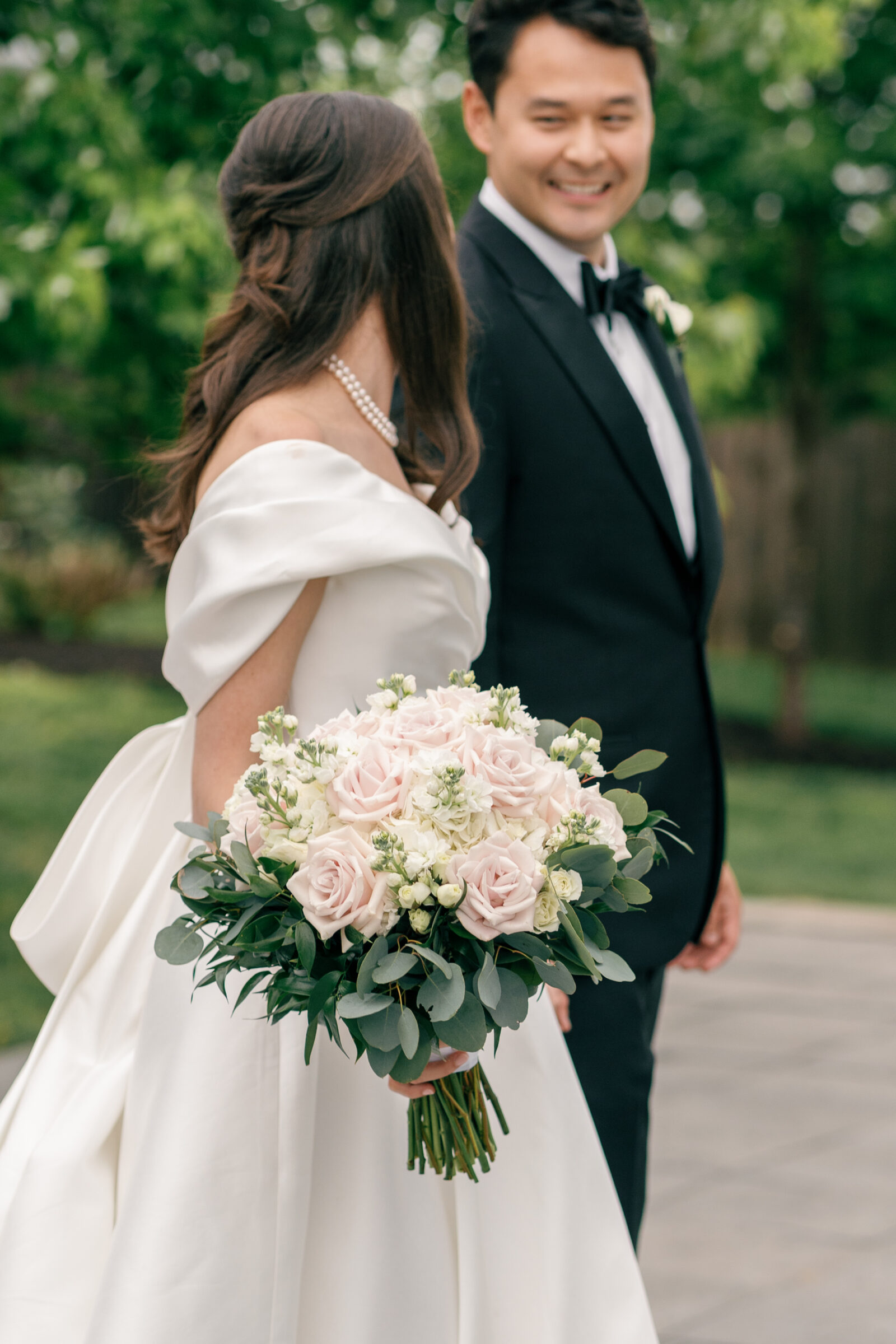 A bride in a satin off-the-shoulder gown holds a lush bouquet of pale pink roses and greenery. She smiles as she walks beside her groom, who is in a classic black tuxedo. The couple enjoys a candid moment, with the groom smiling at her. In the background, greenery and trees add to the outdoor setting.