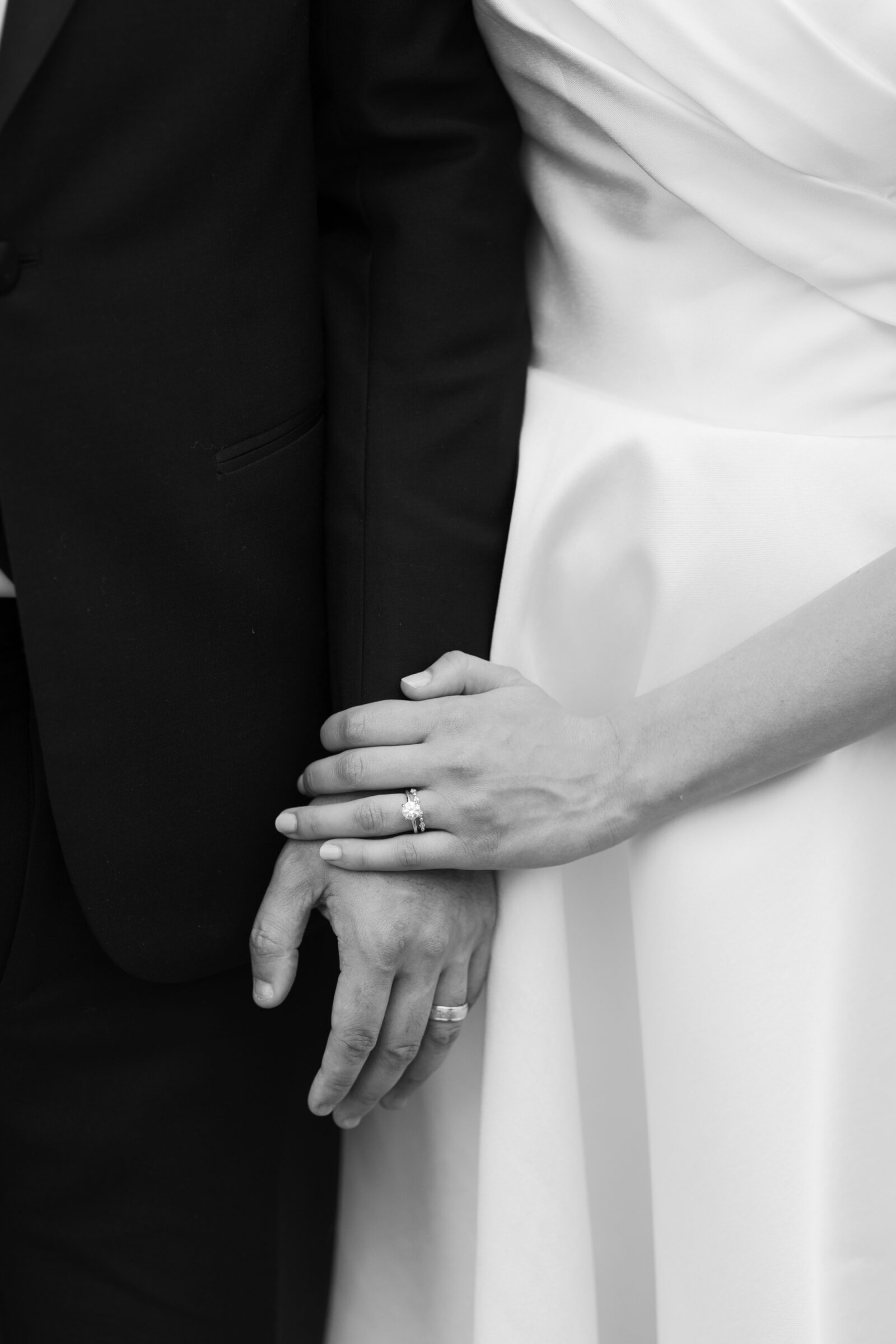 A close-up of the couple’s hands intertwined, displaying their wedding rings. The groom’s hand rests gently on the bride’s, showcasing her engagement ring with the delicate wedding band. The black-and-white photo emphasizes the tenderness of their connection, with the contrast highlighting the rings and their shared bond.
