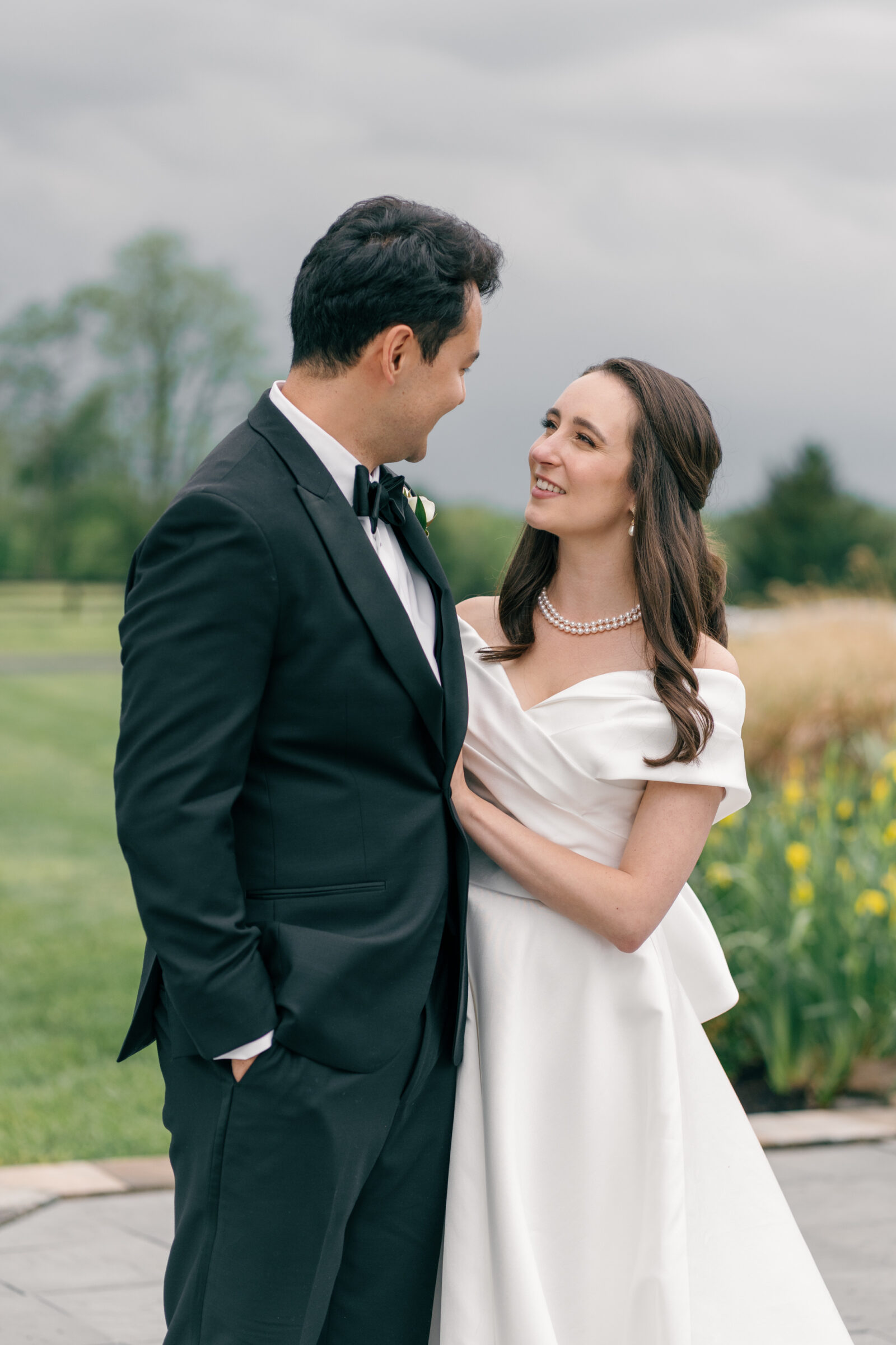 The bride and groom stand close to each other, gazing lovingly into each other's eyes as they smile. The bride wears a pearl necklace, and her satin off-the-shoulder gown flows elegantly around her. The groom, in a classic tuxedo, smiles warmly at her, his hands resting gently at his sides. Behind them, vibrant yellow flowers and green grass provide a beautiful natural backdrop.