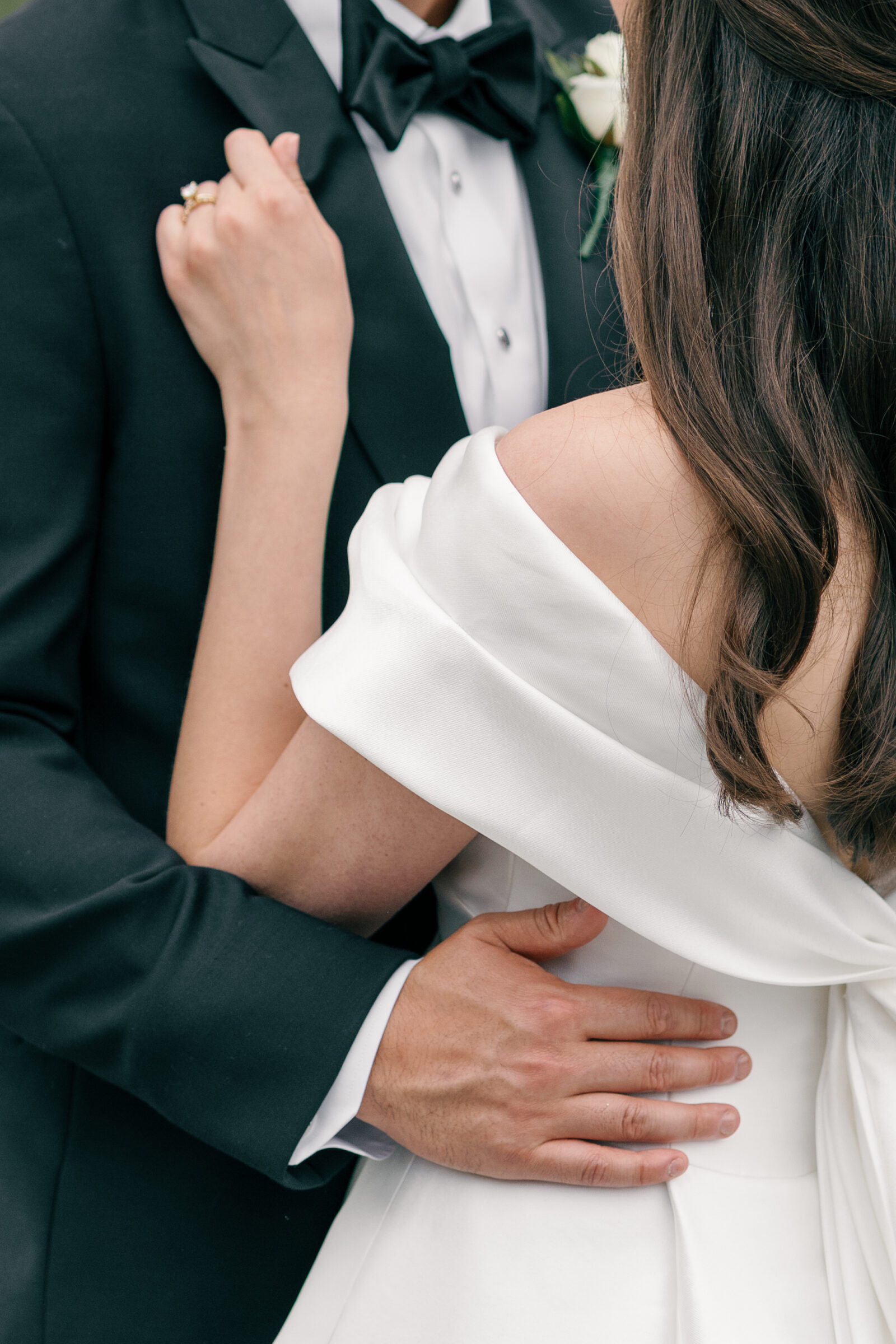 A close-up of the couple in a romantic embrace. The bride, wearing a satin off-the-shoulder gown, holds onto the groom’s tuxedo while his hand gently rests on her waist, showing off her wedding ring. The groom’s hand, elegantly positioned on her back, adds an intimate, tender touch to the moment.