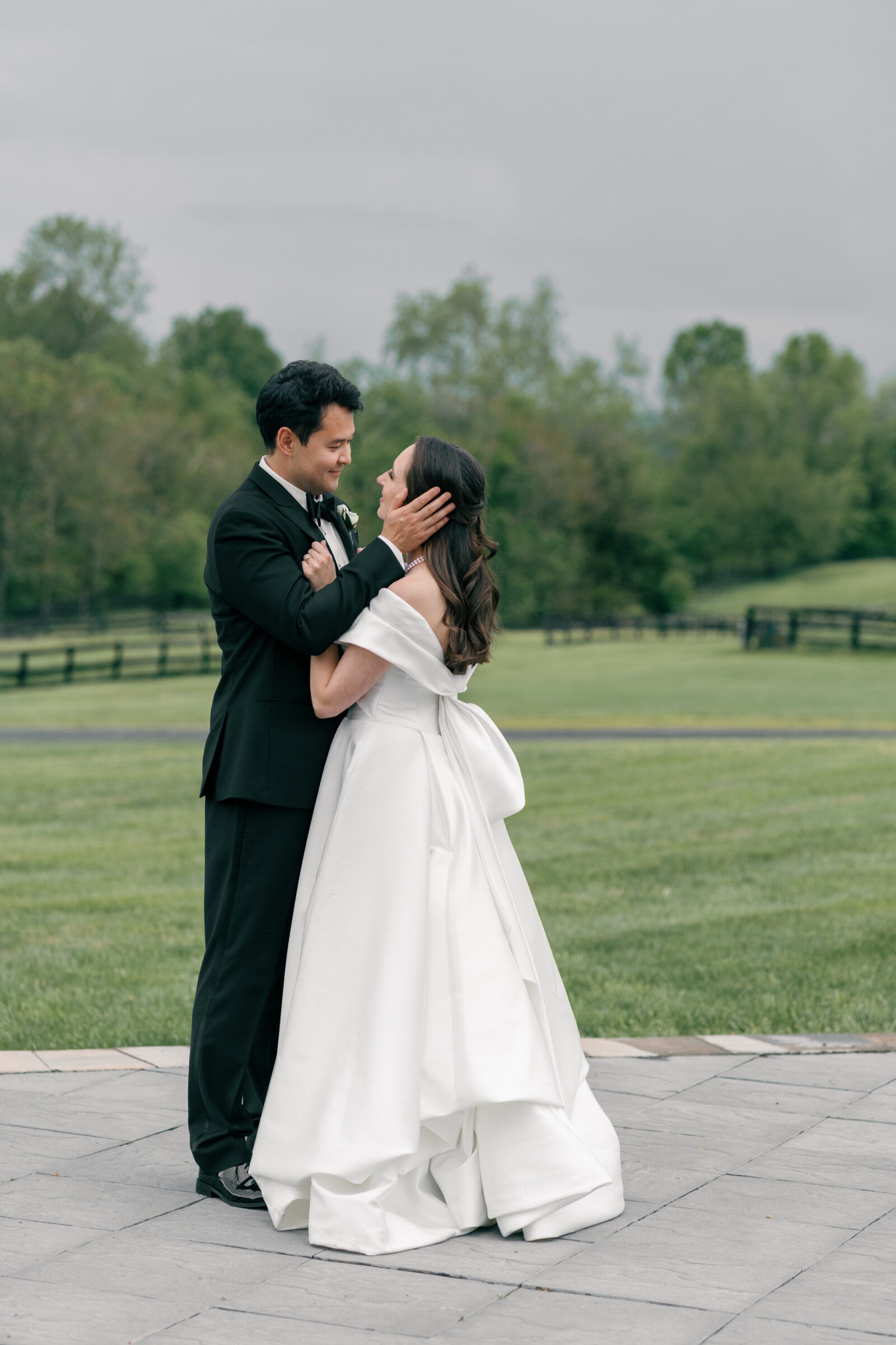 The bride and groom share a tender moment as the groom lovingly holds the bride's face in his hands. The bride smiles up at him with joy, wearing a satin off-the-shoulder gown and a pearl necklace, while the groom, in a classic black tuxedo, gazes at her affectionately. The lush green landscape behind them creates a peaceful, romantic backdrop for this intimate moment.