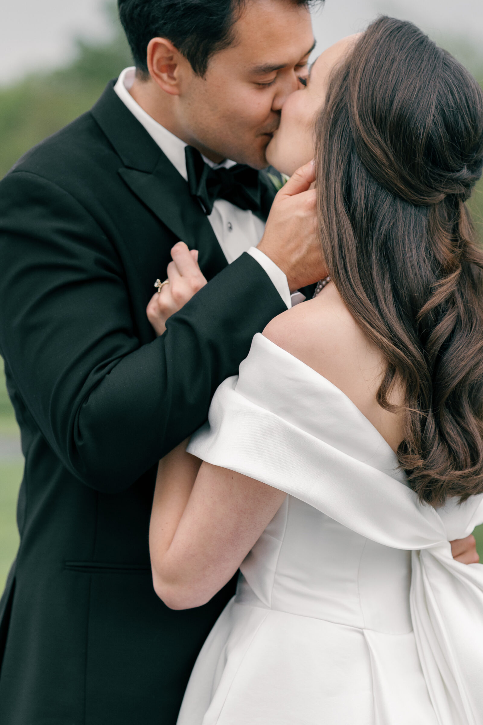 A tender moment between the couple as they share a kiss, standing close together in an intimate embrace. The groom gently holds the bride’s face, while her hand rests on his chest, showcasing her wedding ring. The bride’s off-the-shoulder gown and delicate pearl necklace complement the warmth of their affection.