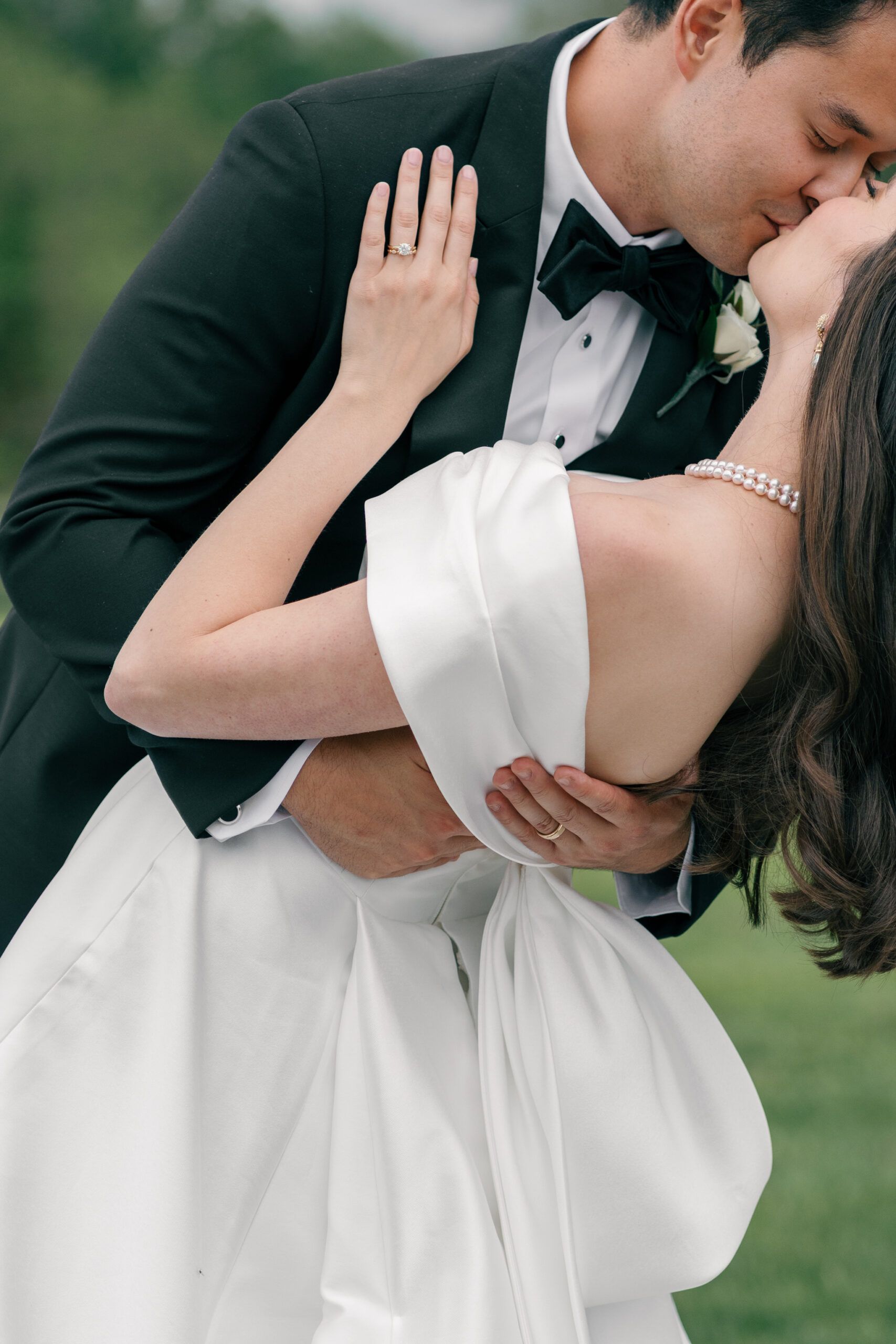 The couple shares an intimate moment, kissing as the groom gently holds the bride while she leans back in a dramatic dip. Her off-the-shoulder satin gown drapes elegantly around her, and her pearl necklace catches the light. The groom’s hand rests on her waist, while her hand is placed on his shoulder, displaying her engagement and wedding rings.