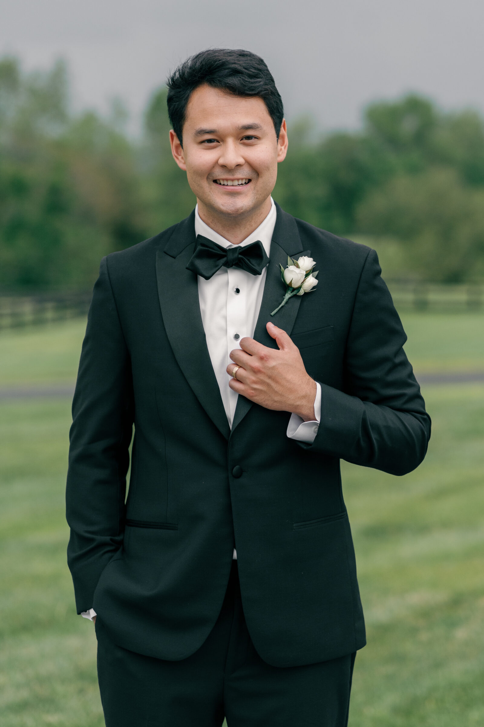 A close-up portrait of the groom, dressed in a classic black tuxedo with a white rose boutonniere. He smiles broadly, adjusting his jacket while his other hand rests casually in his pocket. His confident yet relaxed expression, combined with the lush green background, reflects the joy of the moment.