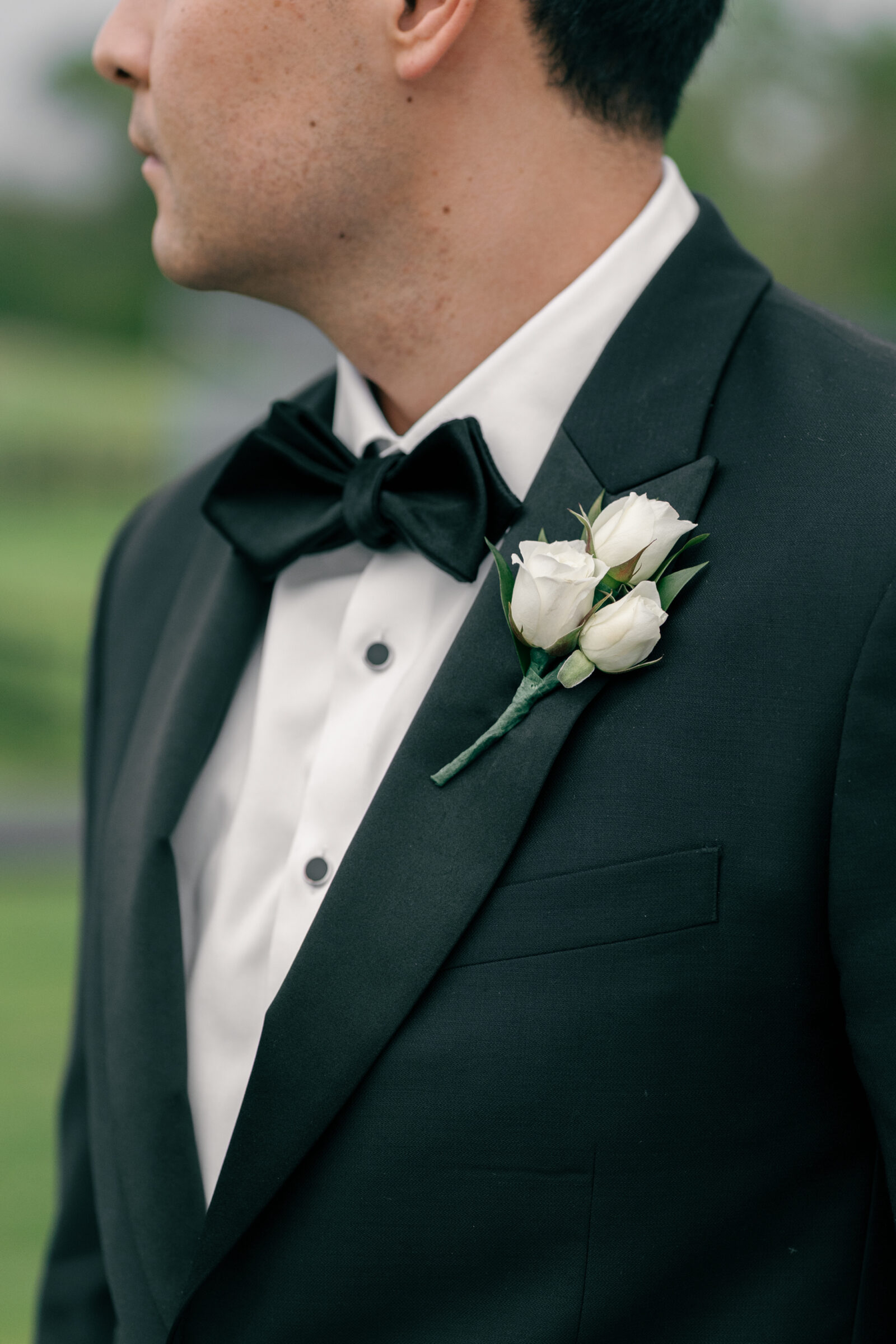 A close-up of the groom's tuxedo with a focus on his bow tie and boutonnière. The black tuxedo jacket is perfectly tailored, with a glossy black bow tie resting neatly on his collar. The boutonnière, consisting of three white roses, is elegantly pinned to his lapel, contrasting beautifully with the dark jacket and setting a refined tone for the wedding day.