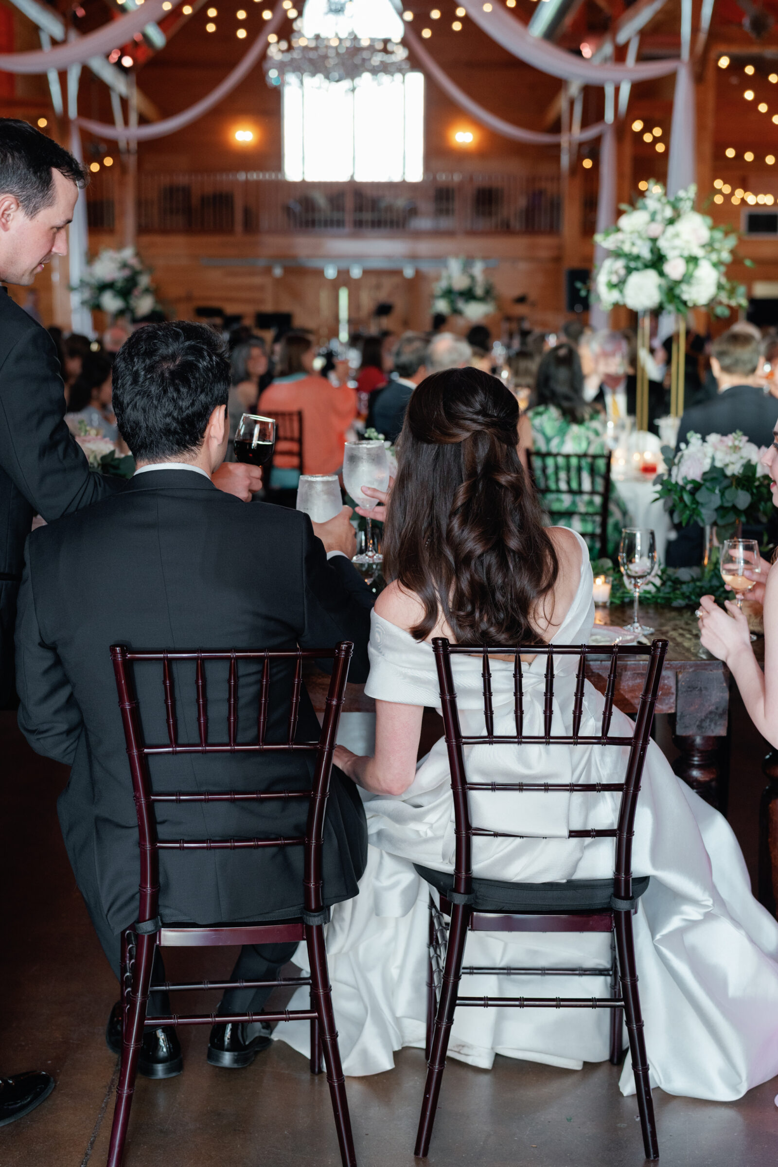 A back view of the bride and groom sitting at their wedding reception table. They are facing a beautifully decorated venue, with elegant chandeliers, large floral arrangements, and guests enjoying the evening. The bride’s gown cascades elegantly behind her, and the groom is holding a glass of wine. The couple enjoys a quiet moment together amidst the lively atmosphere of their celebration.