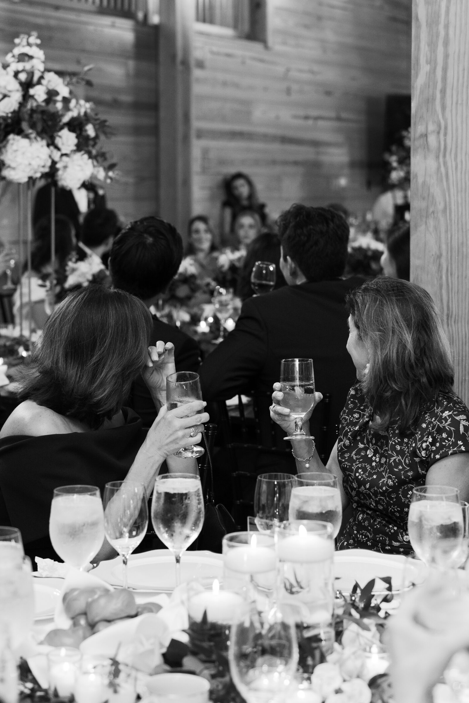 A black and white photograph of guests at a wedding reception, seated at their tables with glasses in hand. In the foreground, two guests are raising their glasses, one wiping away a tear. The tables are beautifully decorated with candles, floral arrangements, and glasses of water. The background shows more guests listening to a speech, with the warm, inviting atmosphere of the barn venue in the background.