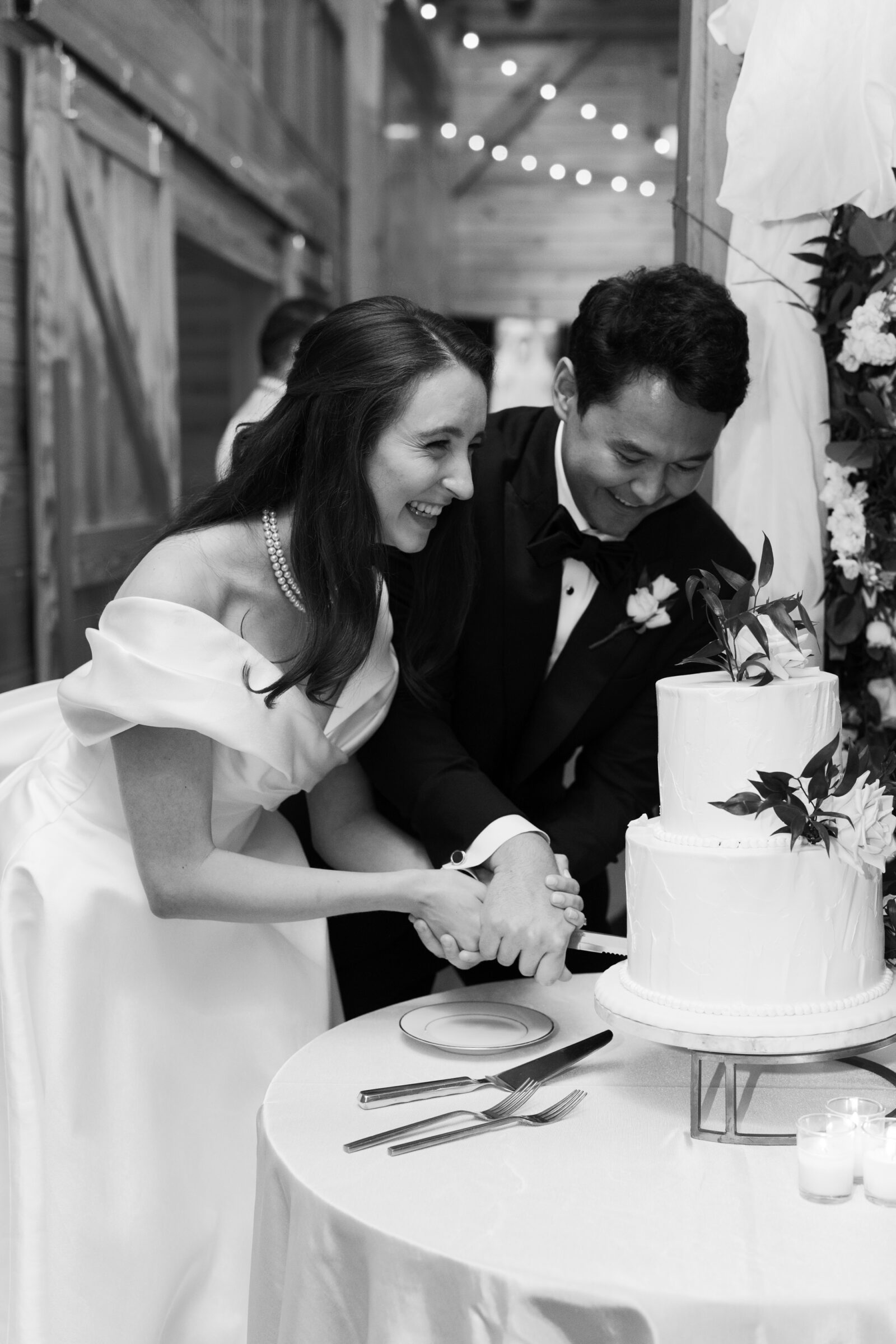 A black-and-white photo of the bride and groom cutting their wedding cake together. Both are smiling, with the bride holding the cake knife in her hands and the groom gently guiding her. The cake is a two-tiered white design, adorned with delicate greenery. The scene is set at a table with a plate and utensils nearby, and soft candlelight adds a warm glow to the ambiance.