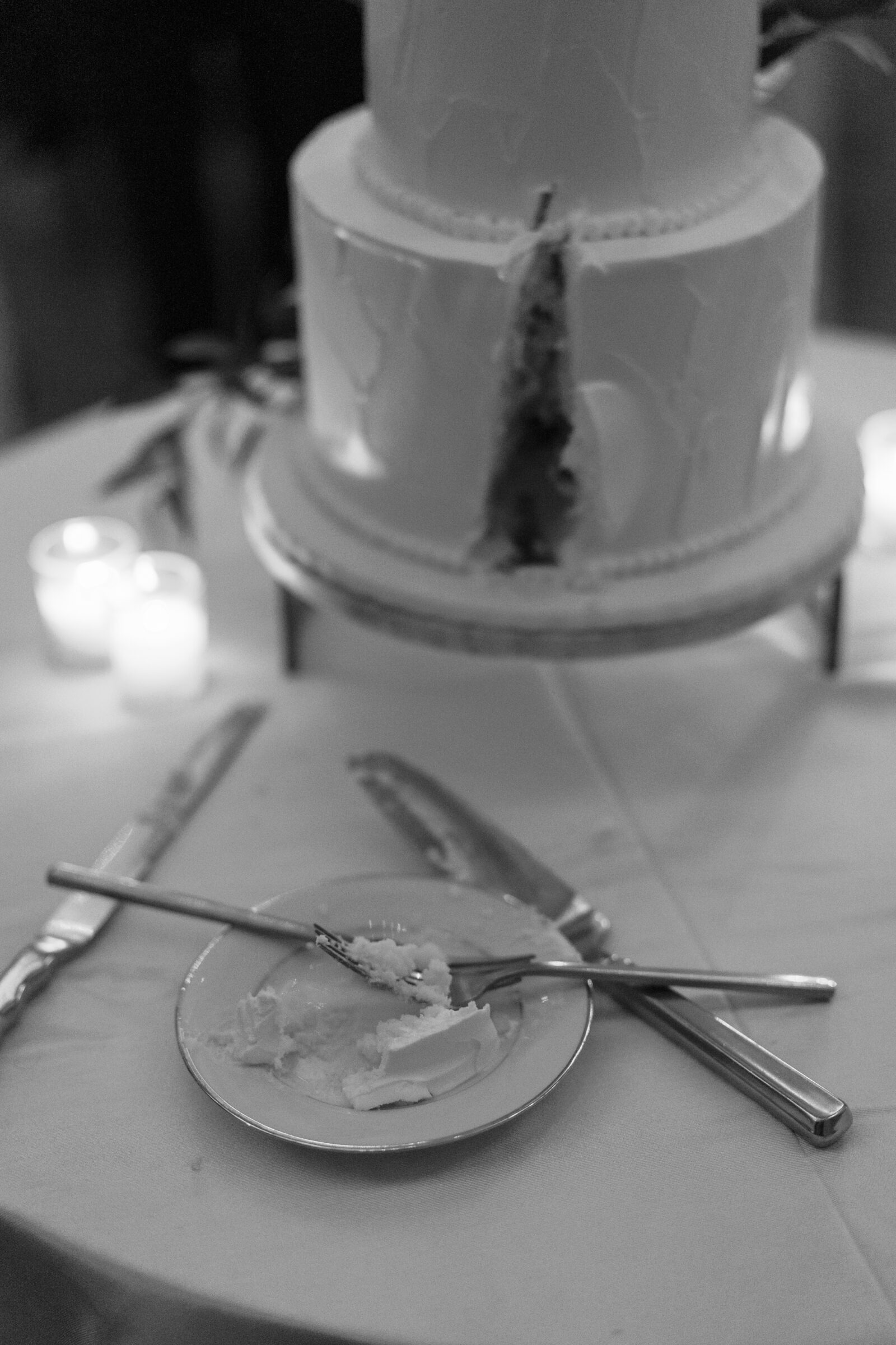 A black-and-white close-up of a wedding cake, partially sliced, with a plate of cake on the table. The plate contains a small portion of cake with a fork and knife beside it. The cake is decorated with smooth white icing and placed on a silver cake stand, with soft candlelight glowing in the background.