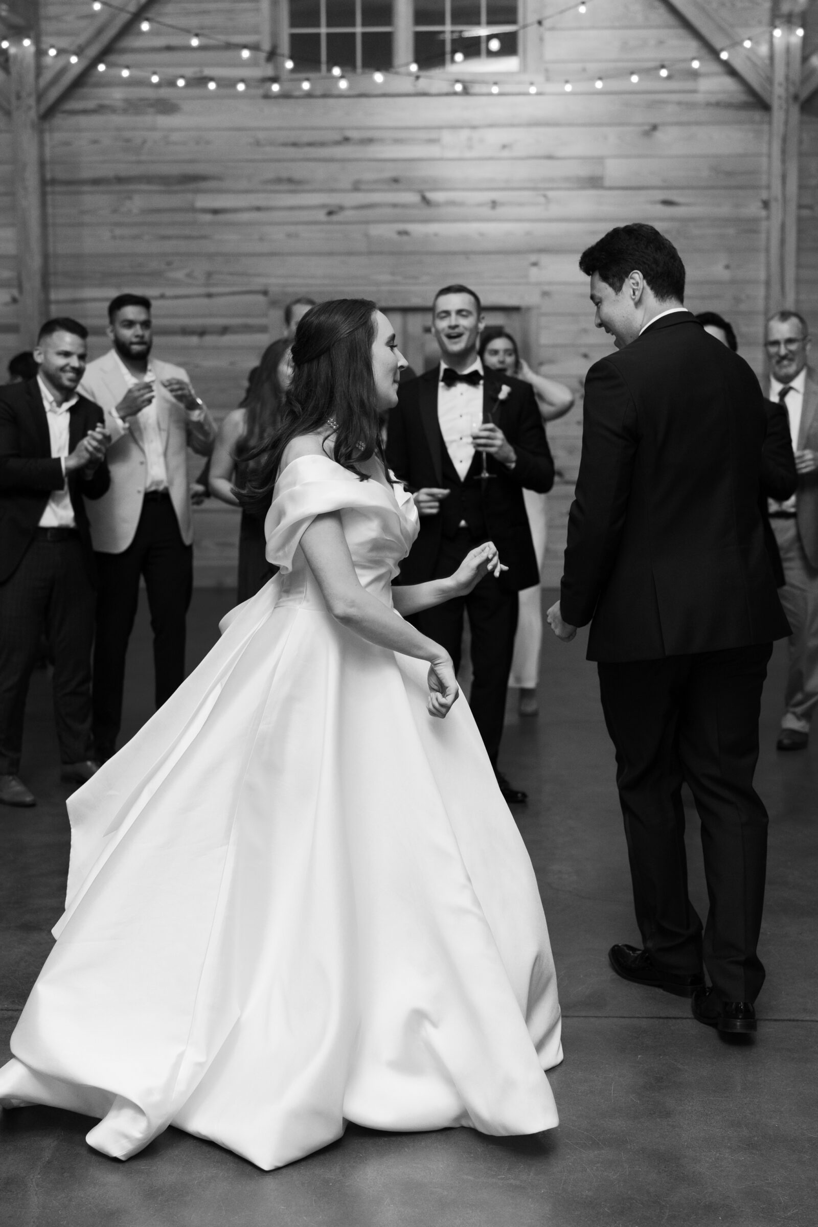 A black-and-white photo of the newlywed couple sharing a joyful moment dancing at their wedding reception. The bride, wearing a beautiful off-the-shoulder gown, twirls as she shares a smile with her groom, who is dressed in a tuxedo. Guests are gathered around, clapping and celebrating in the background under the warm glow of string lights in the barn venue.