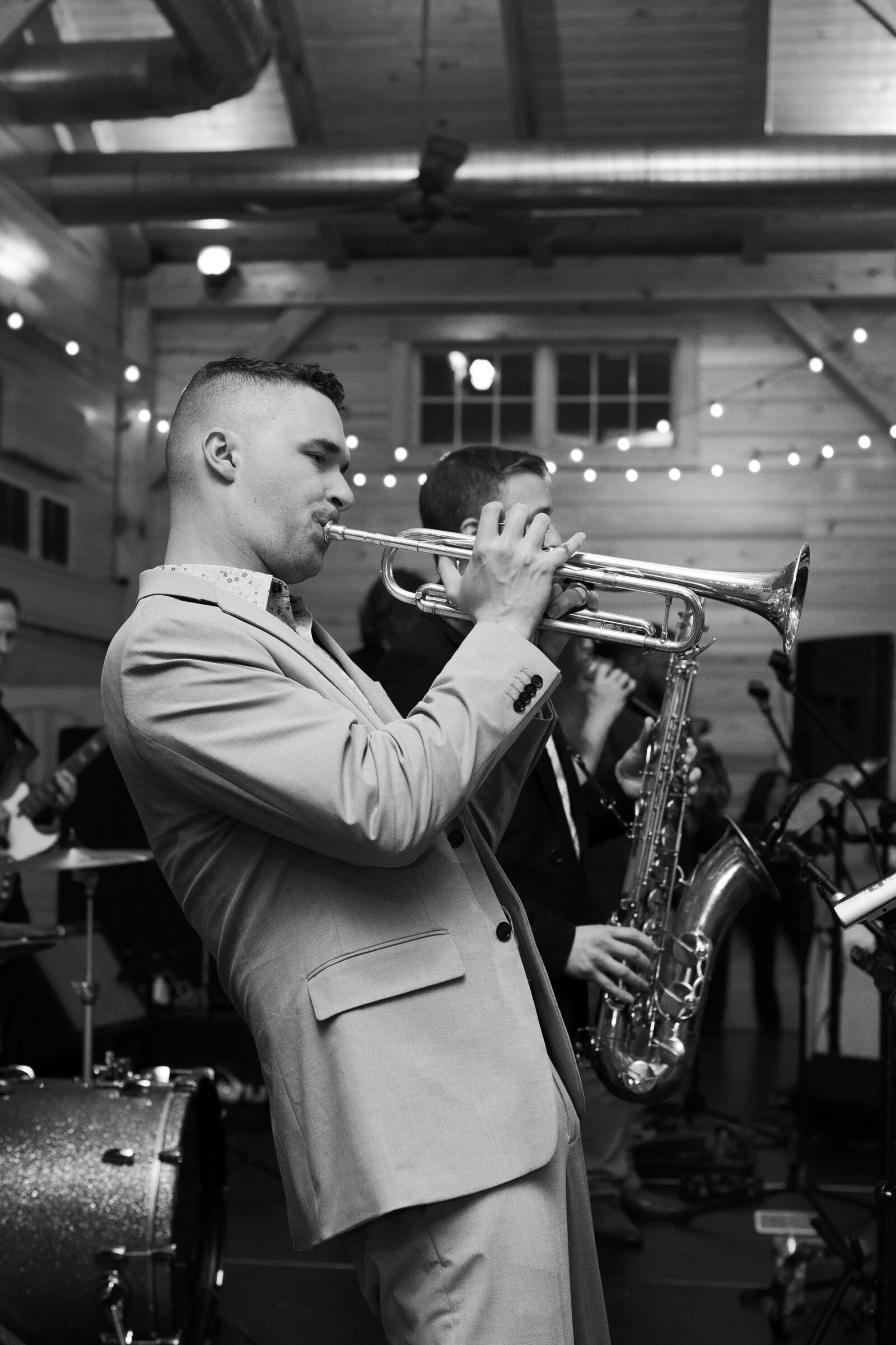 A black-and-white photo of a trumpet player performing at the wedding reception. He is dressed in a light-colored suit, passionately playing his instrument, with a saxophone player visible in the background. The atmosphere is lively, with soft string lights illuminating the barn-like venue.