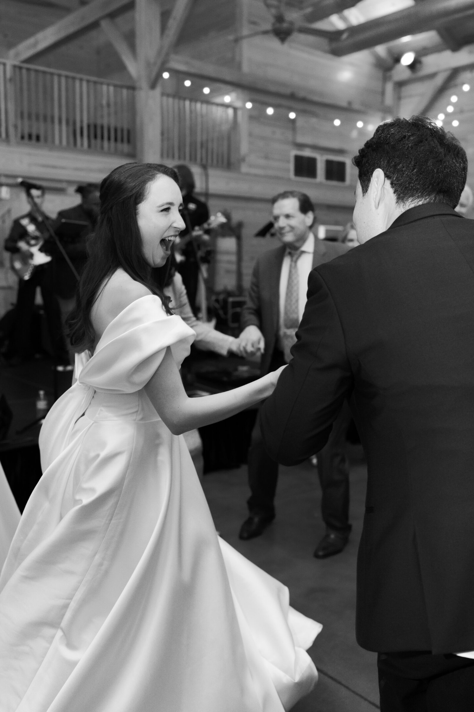 A black-and-white photo of the bride and groom joyfully dancing together at their wedding reception. The bride, dressed in her elegant off-the-shoulder gown, smiles widely as she laughs and holds her husband's hand. The groom, in a tuxedo, shares in the joyful moment. Guests are gathered around in the background, celebrating, as the live band plays in the distance under string lights in the rustic barn venue.