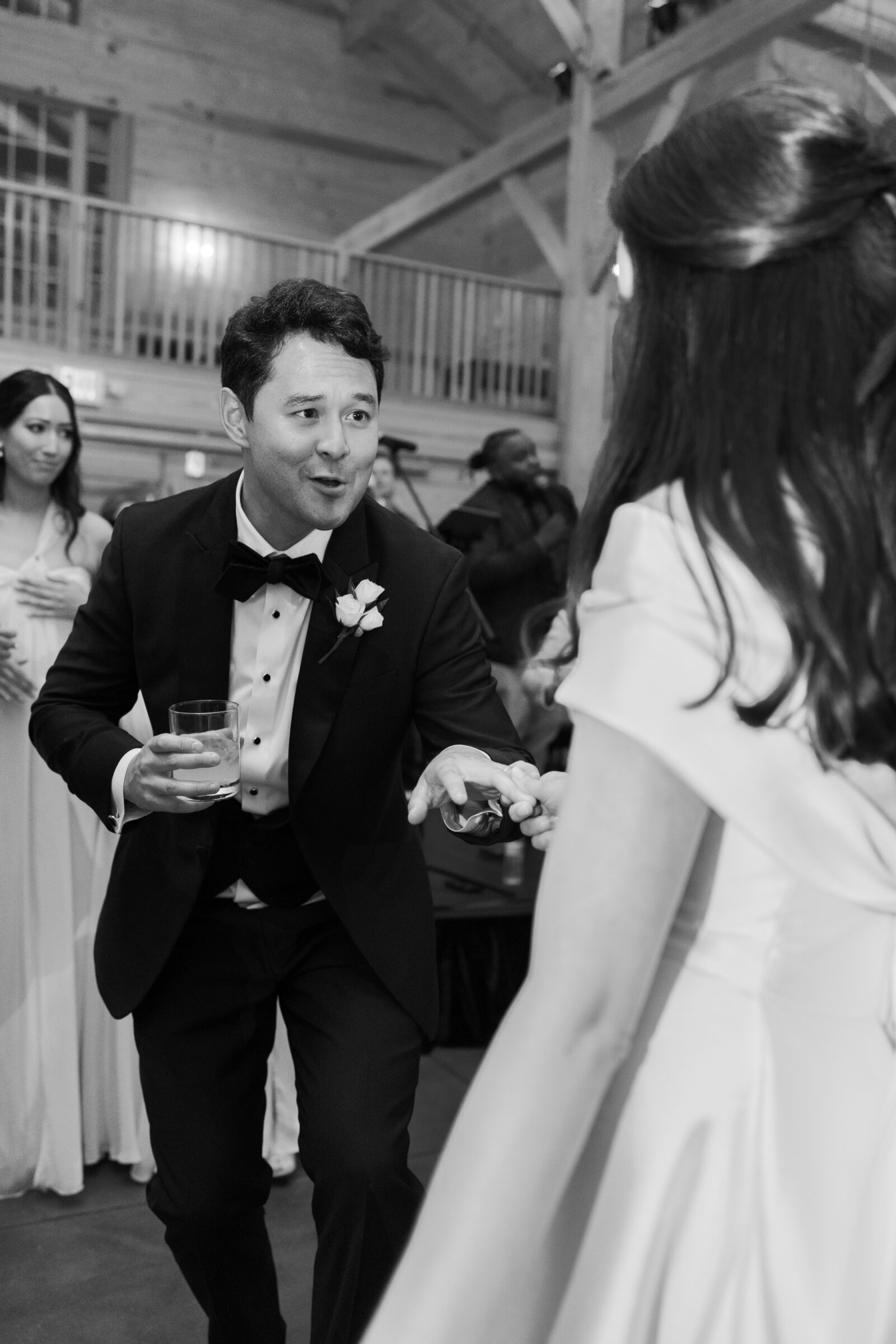 A black-and-white photo of the groom, wearing a tuxedo, extending his hand toward the bride with a playful expression as they dance during their wedding reception. He holds a drink in his other hand. The bride, in an off-the-shoulder wedding gown, reaches out to him. Guests are visible in the background, enjoying the celebration in the rustic barn venue.