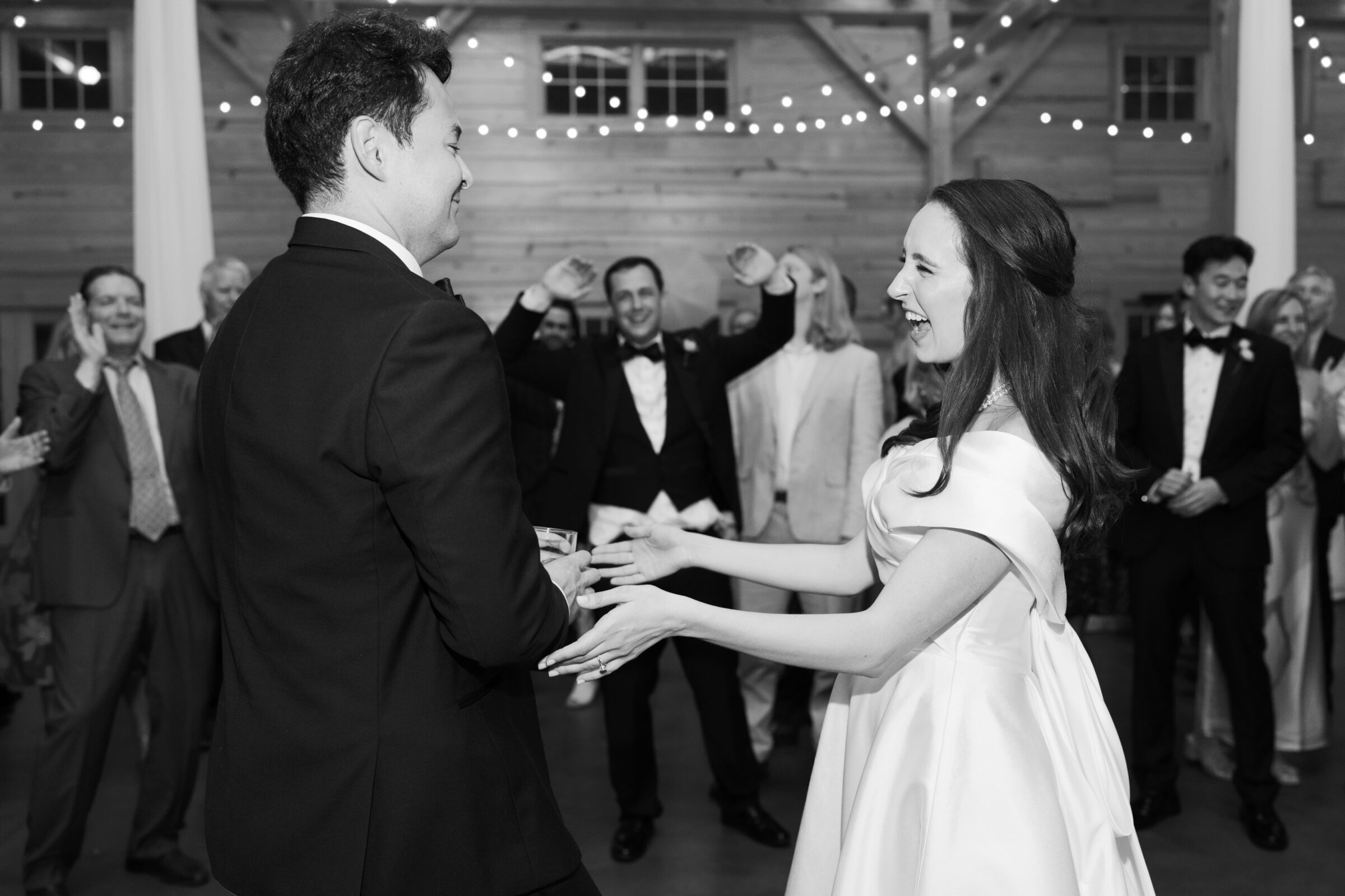 A black-and-white photo of the bride and groom laughing and holding hands as they dance during their wedding reception. The groom, wearing a tuxedo, holds a drink in his other hand. Guests in the background are cheering and raising their arms, enjoying the moment in the lively barn venue, decorated with string lights.