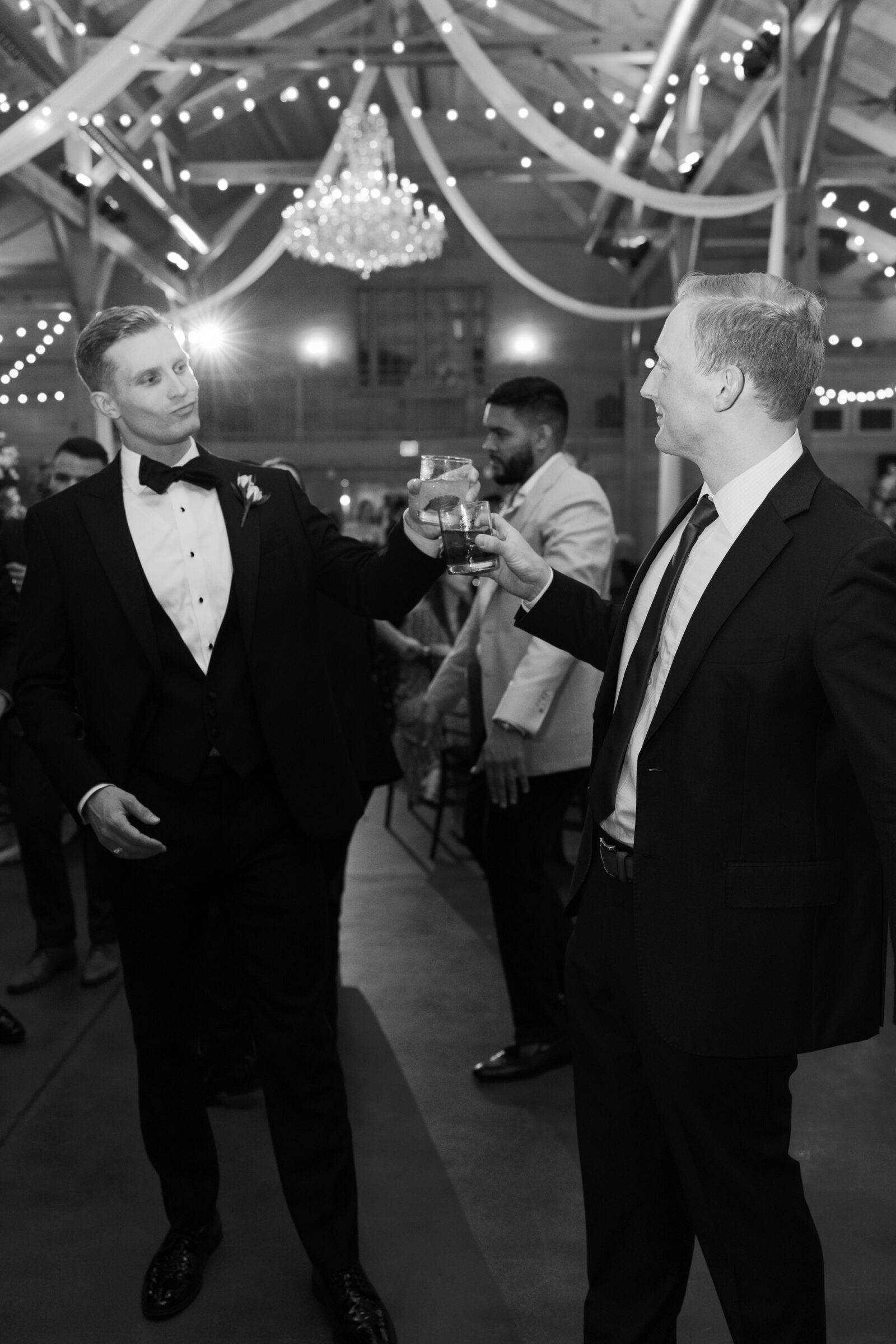 A black-and-white photo of two male wedding guests toasting glasses on the dance floor. Both are dressed in tuxedos or suits, one wearing a bow tie and the other a tie. The background shows other guests, and the setting is a festive wedding reception with string lights and a large chandelier overhead. The atmosphere is lively, with the guests enjoying the celebration.