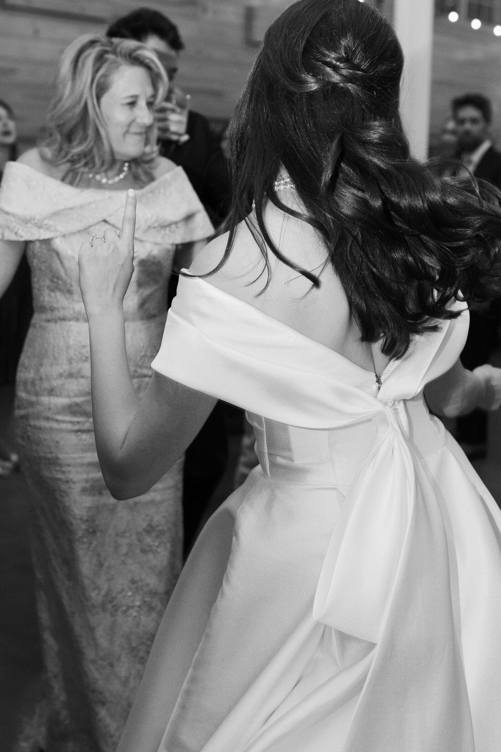 A black-and-white photo of the bride dancing at her wedding reception, seen from the back. She wears a stunning off-shoulder dress with a large bow at the back of her gown. She gestures with one hand raised in the air while dancing with friends. In the background, a woman in a sparkling gown is also dancing, and other guests enjoy the lively atmosphere of the celebration.